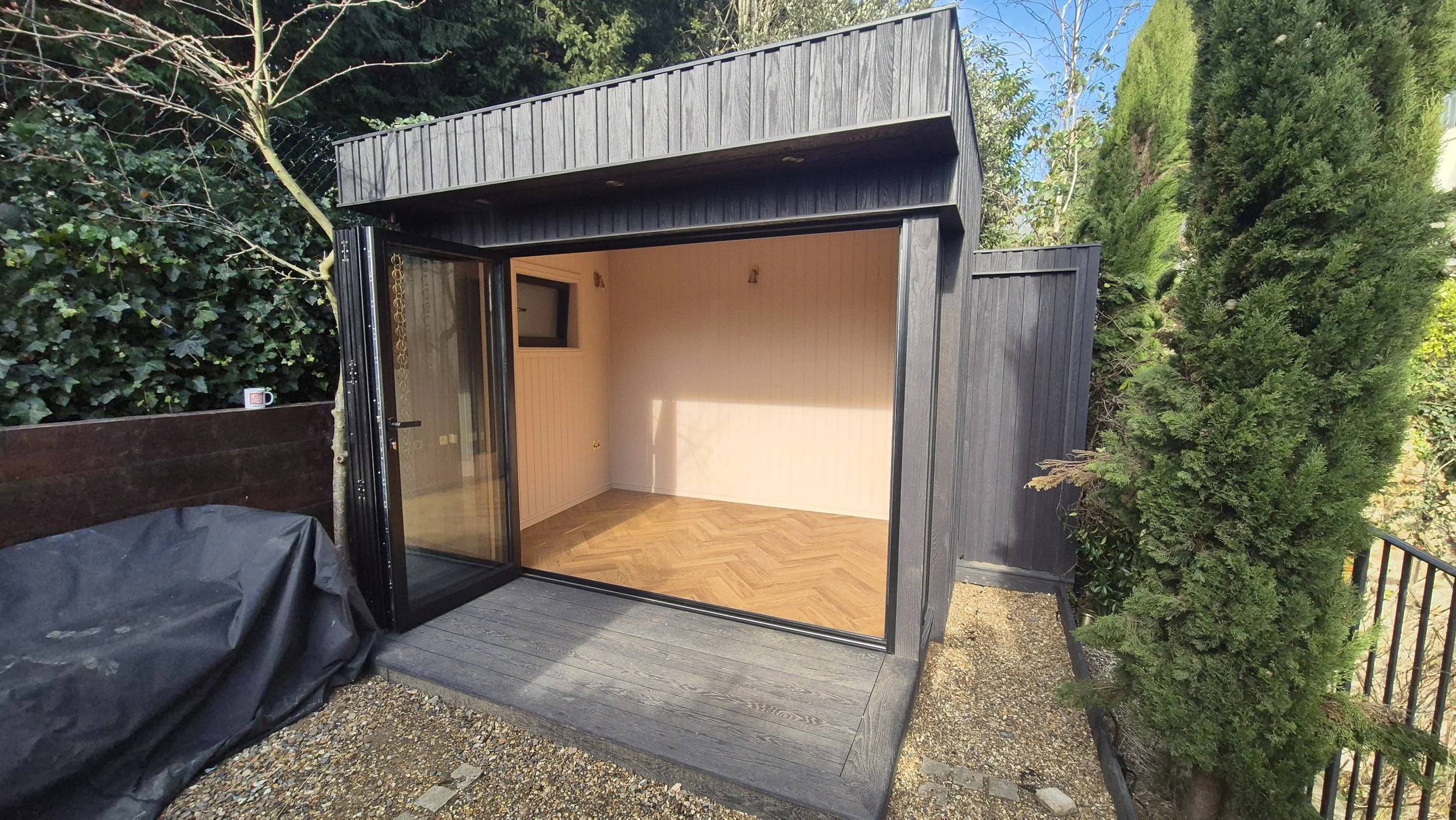Small modern black wooden outdoor structure with large glass doors opening to a room with light-colored walls and wood flooring, surrounded by greenery and shrubs.
