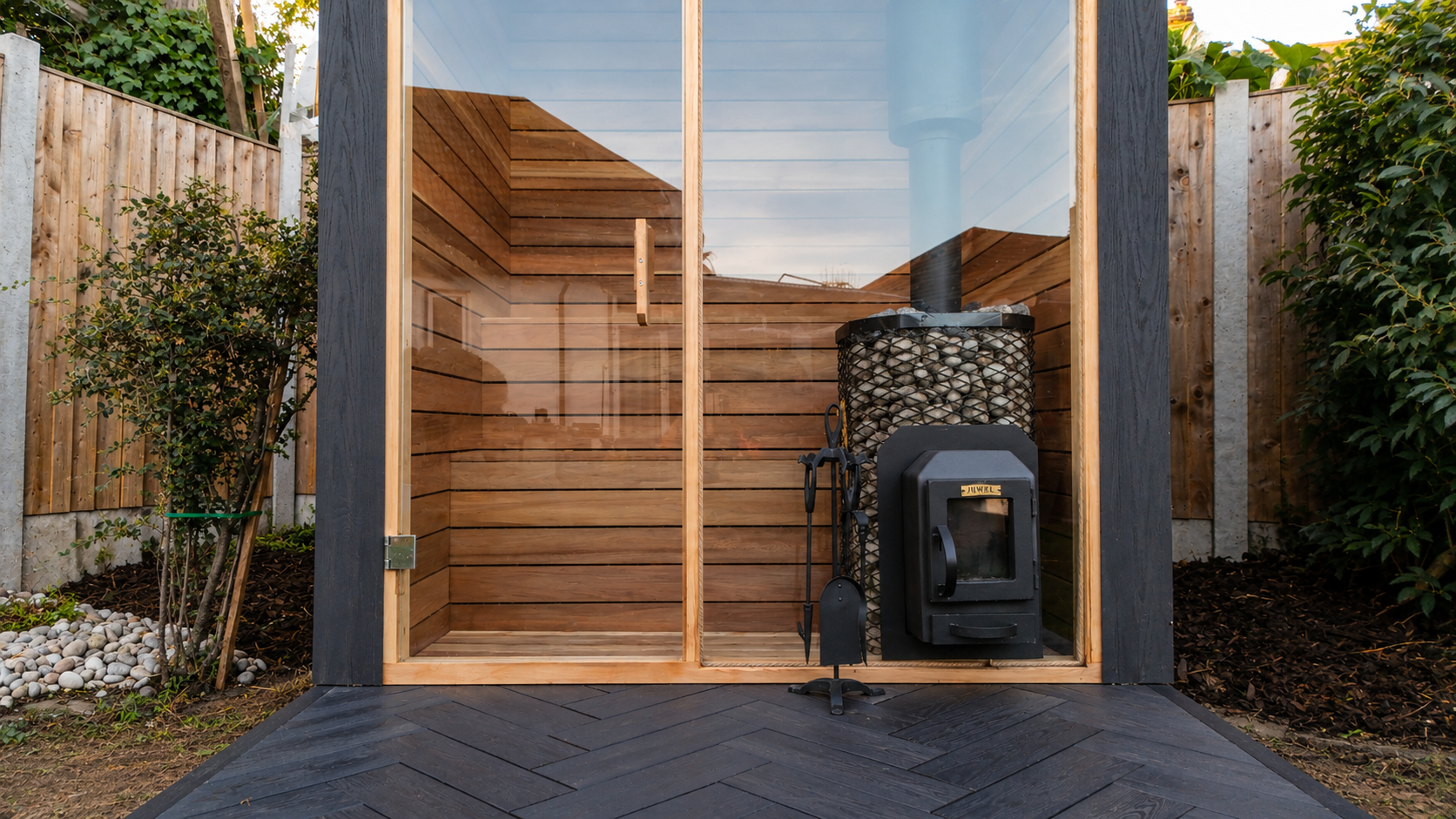 Outdoor sauna with wooden interior and charcoal stove, enclosed by glass and surrounded by landscaped garden.