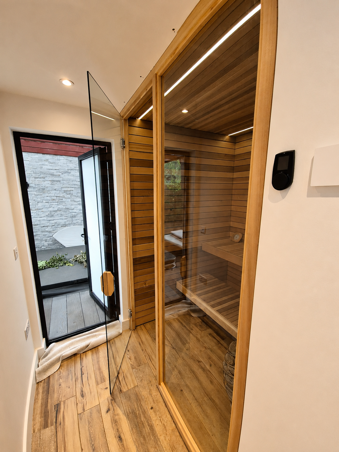 Interior view of a wooden sauna with glass door, adjacent to a porch area with stone wall and outdoor bathtub.