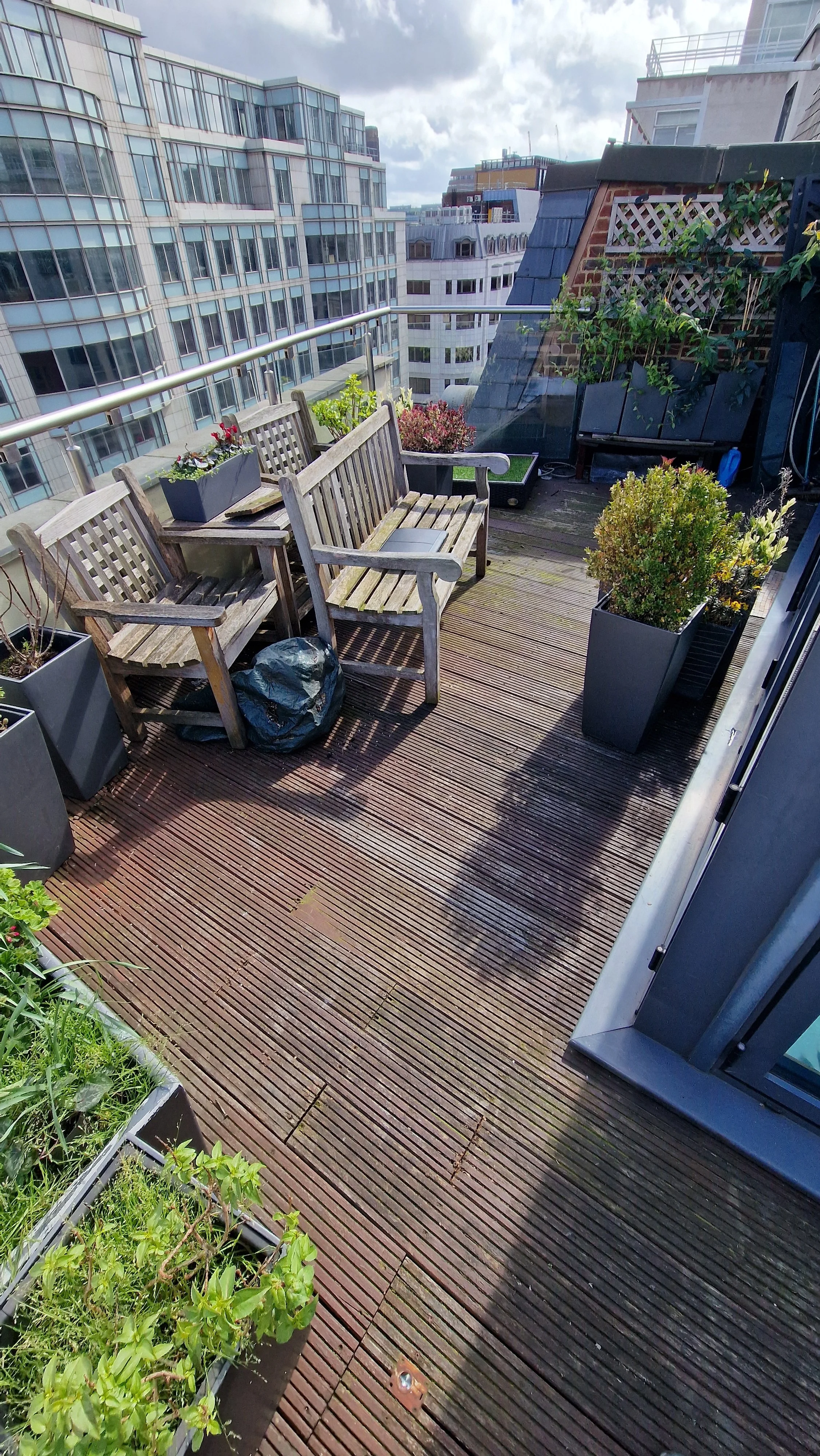 A rooftop balcony with wooden decking, potted plants, and outdoor furniture including two wooden benches and a small table. The background shows neighboring tall buildings and partly cloudy sky.