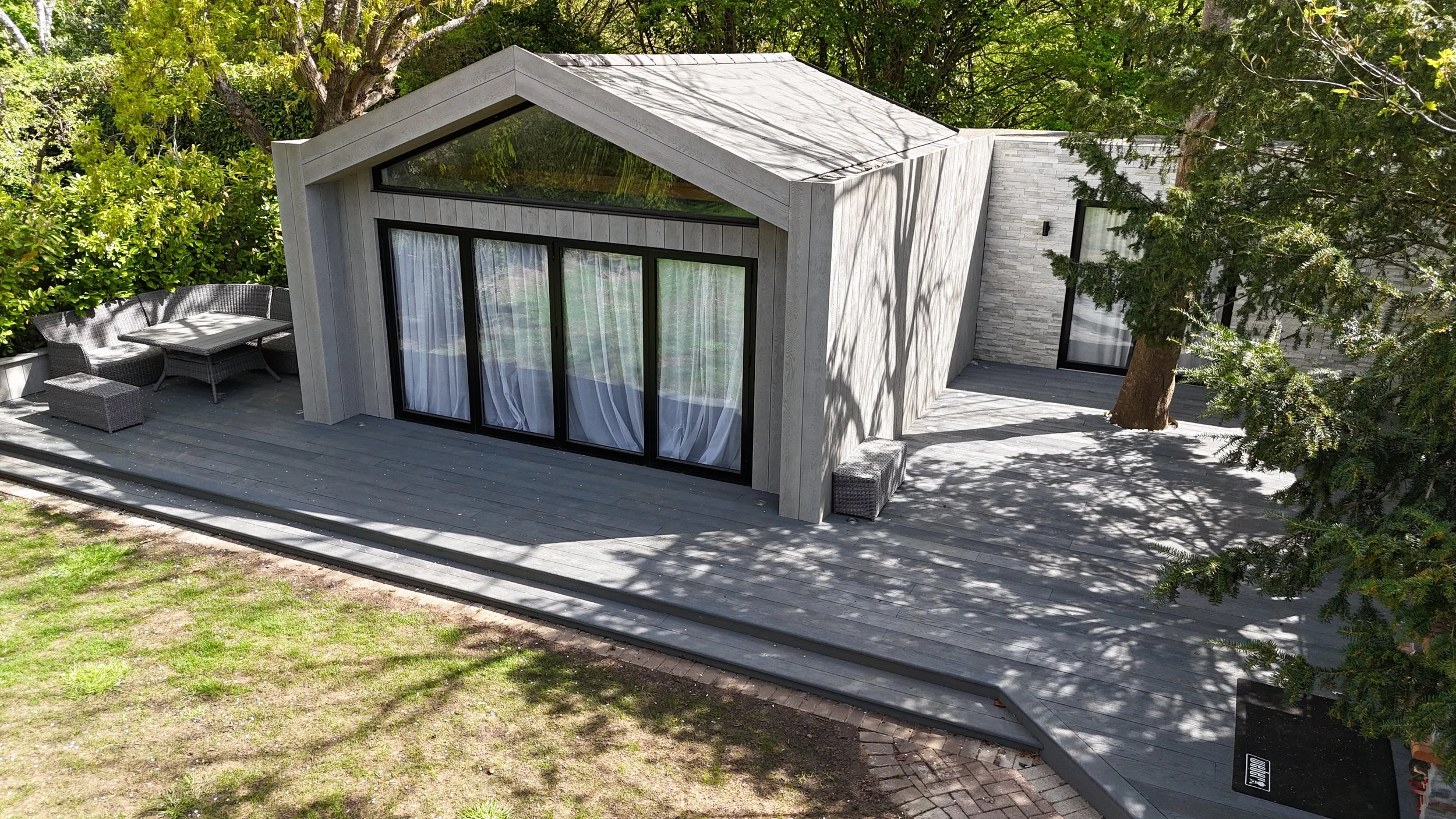 A modern house with a gray exterior, large glass sliding doors, and a deck surrounded by trees and green foliage.