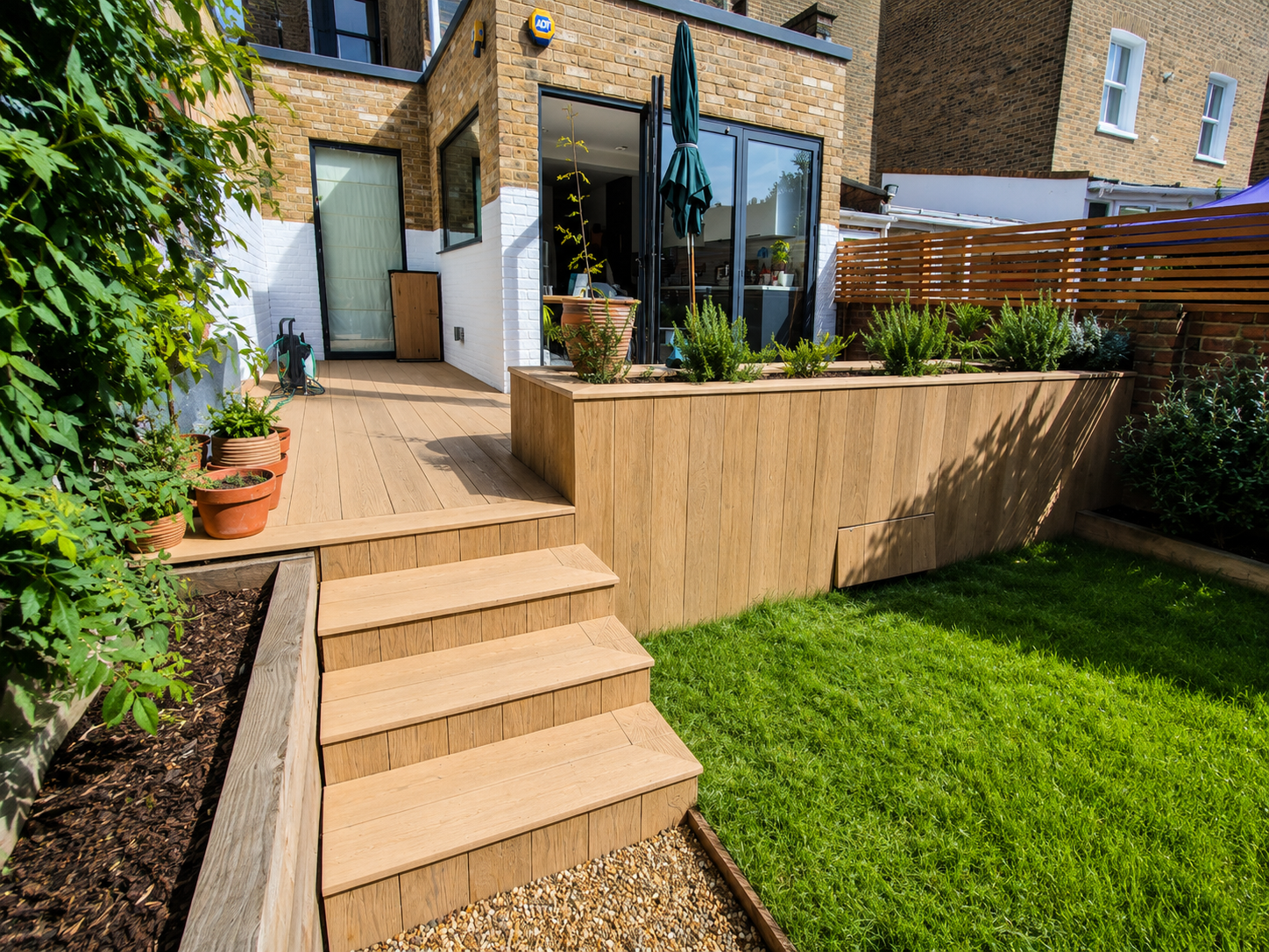 A backyard patio with wooden steps leading up to a patio area with plants, a large potted tree, and a glass door opening into the house. There is green grass in the foreground, and a garden bed with potted plants and shrubs on the right side.