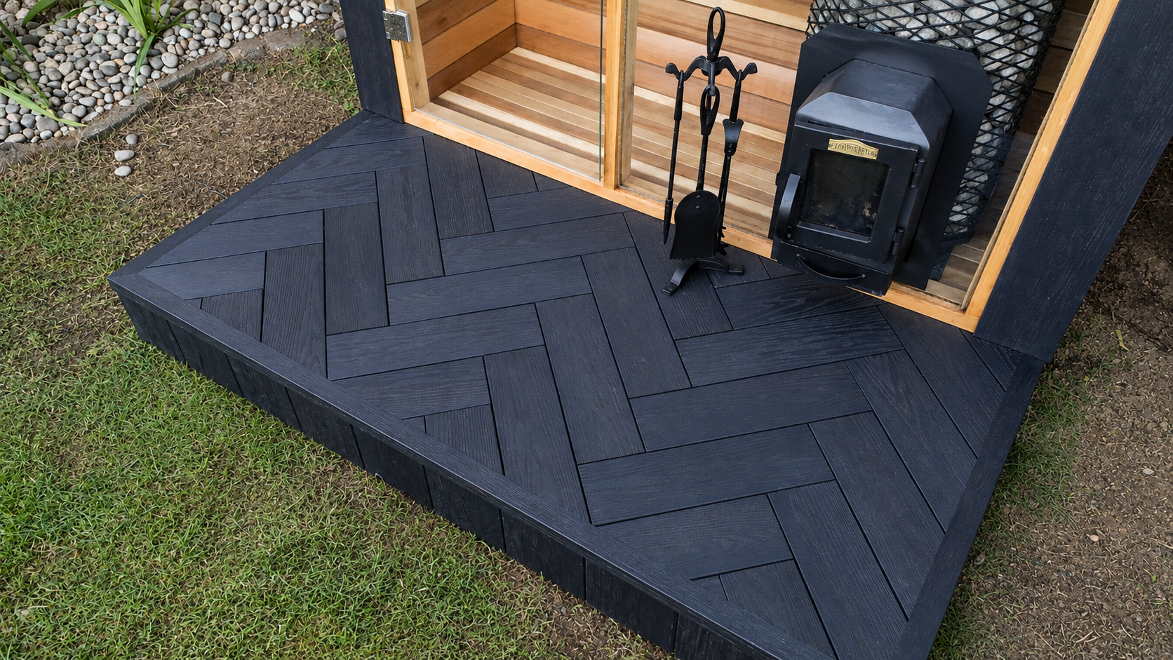 Black wooden deck with a set of fireplace tools, a wood stove, and a screened enclosure with a wooden interior. Surrounding the deck is grass and gravel with small rocks.