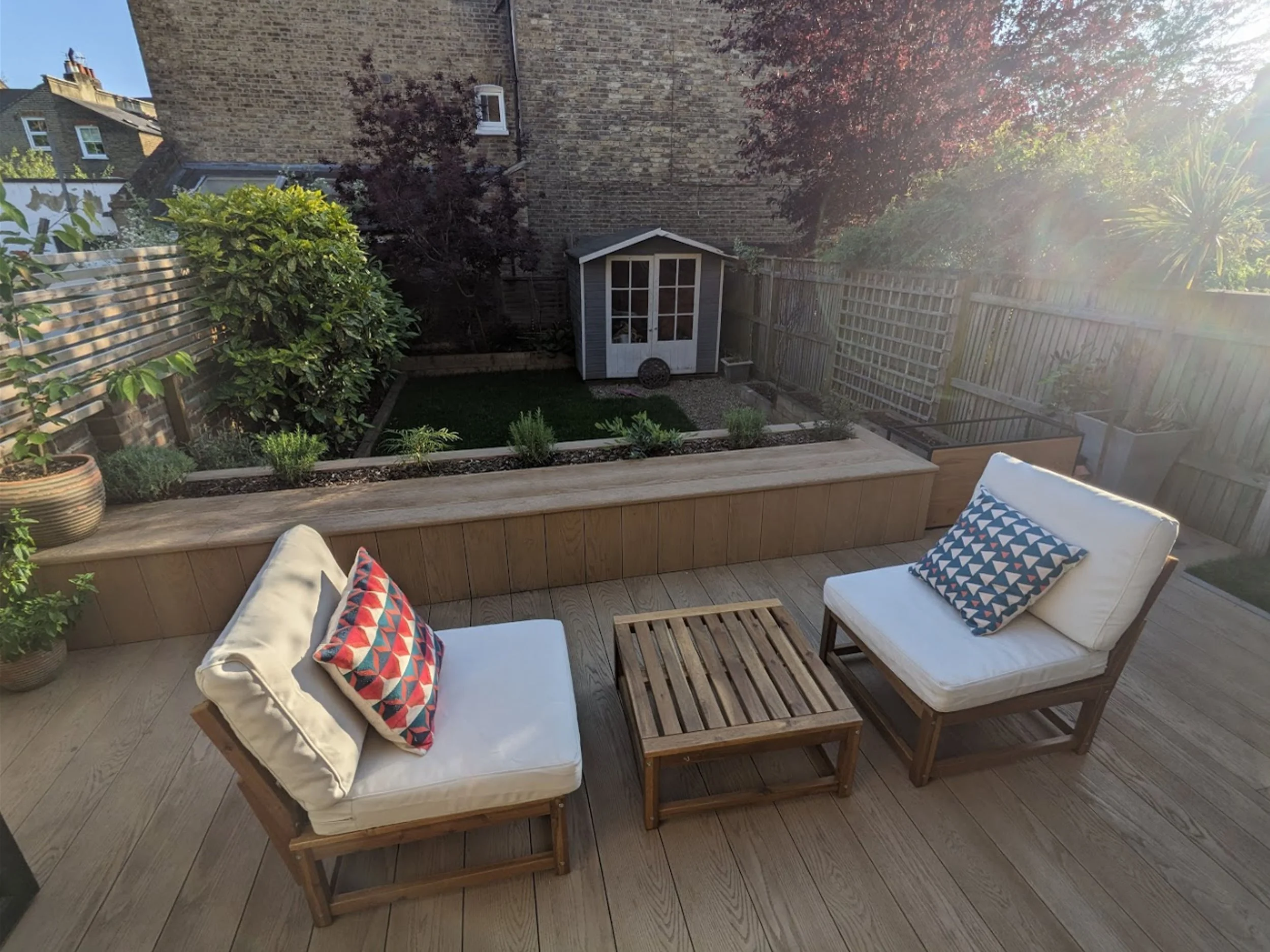 A backyard patio with outdoor furniture including a beige cushioned armchair, a wooden coffee table with a slatted top, and a wooden sofa with beige cushions, decorated with colorful patterned throw pillows. The patio is bordered by wooden planters with green plants, and a low wood railing. In the background, there is a small garden shed, a grassy area, and a wooden fence with trees and bushes nearby. The sun is shining brightly, creating a glare on the right side of the image.