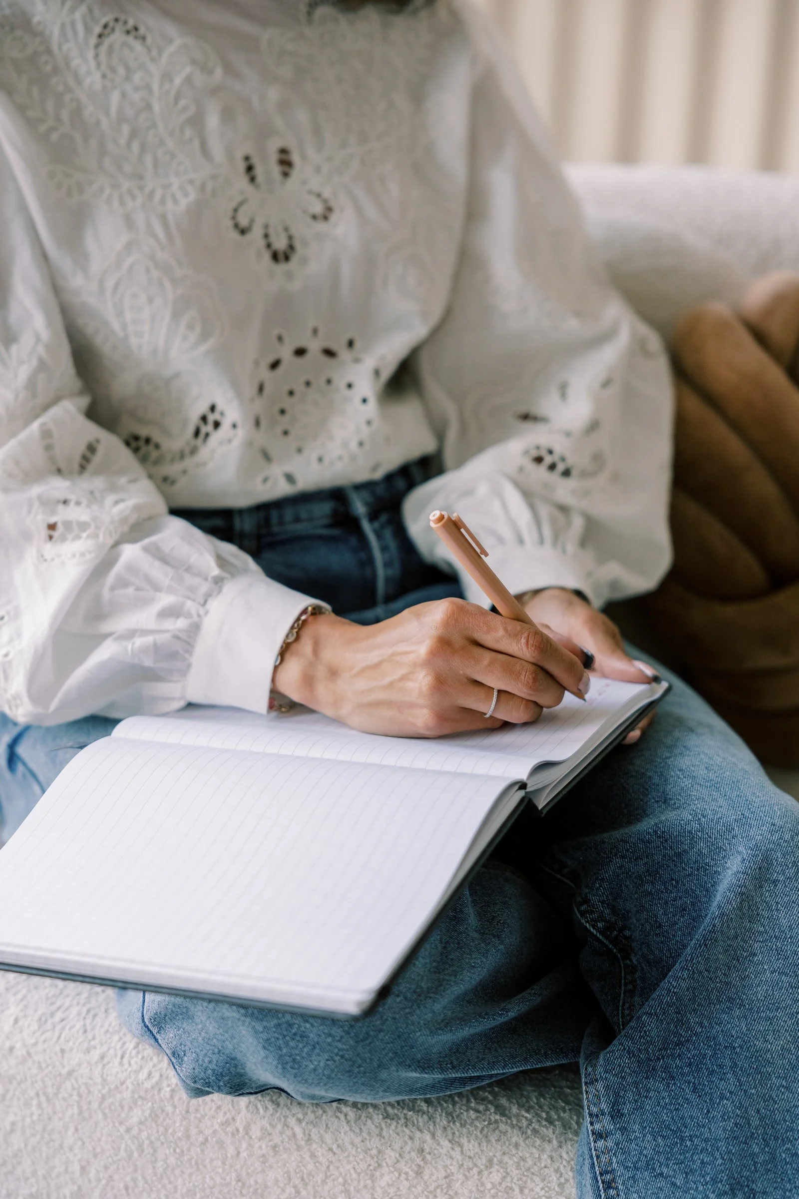 A person sitting on a beige carpet floor, writing in an open lined notebook with a pink pen, wearing a white embroidered blouse and blue jeans.