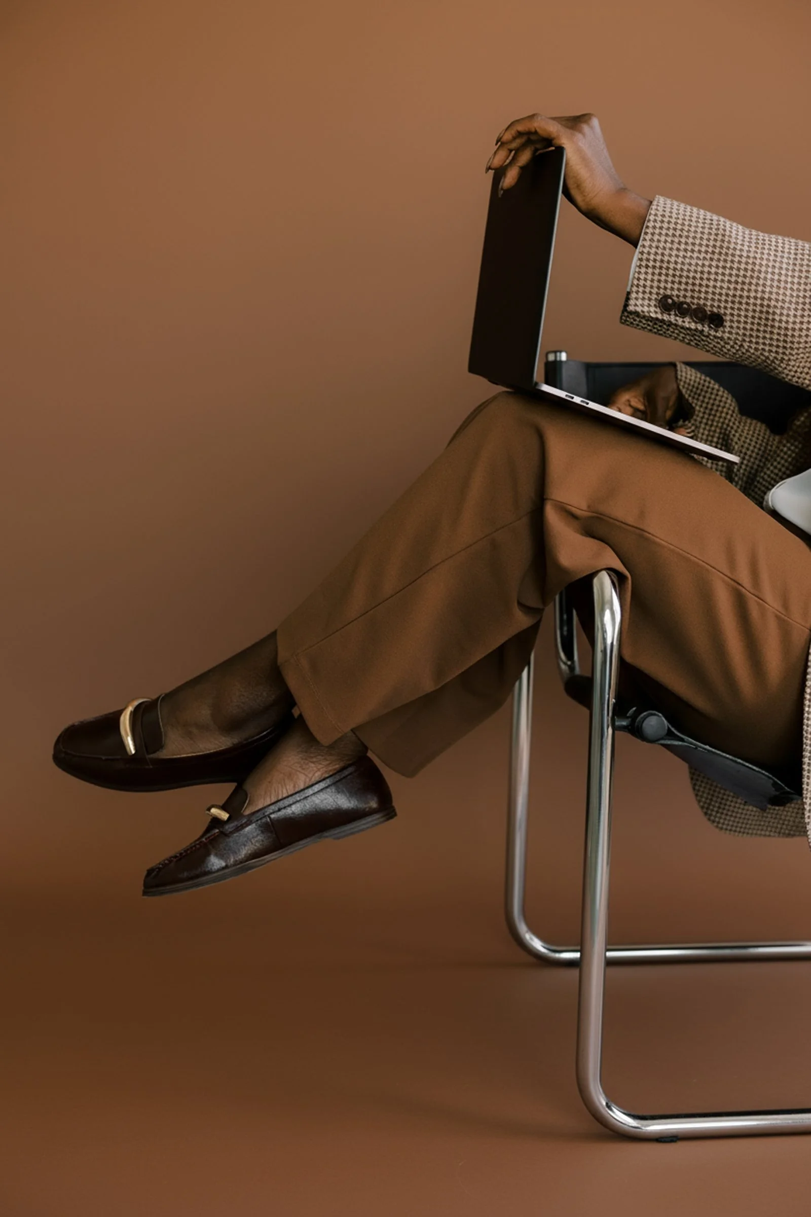 A person sitting on a chair wearing brown dress shoes and brown trousers, with a brown patterned blazer, holding a laptop with a brown background.
