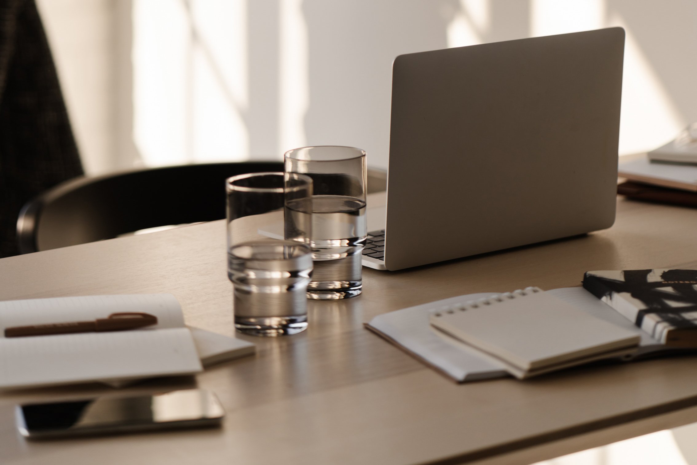 A wooden desk with a closed silver laptop, two glasses of water, notebooks, and a pen in bright natural light.