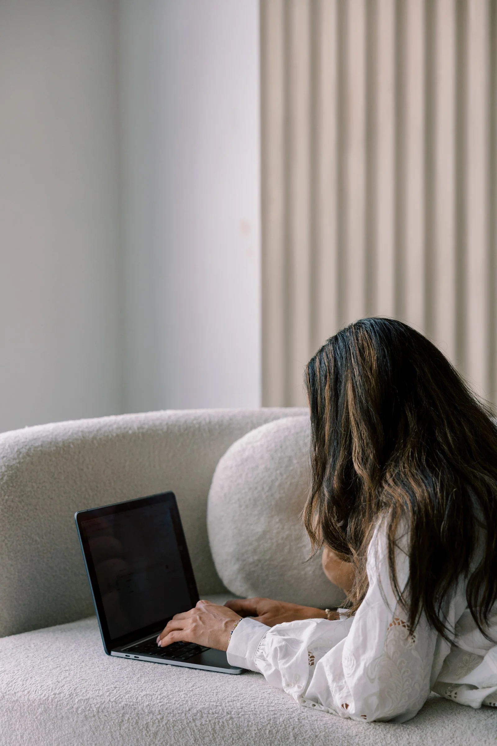 A woman with brown hair working on a laptop, sitting on a cream-colored couch in a room with cream-colored walls and vertically striped curtains.