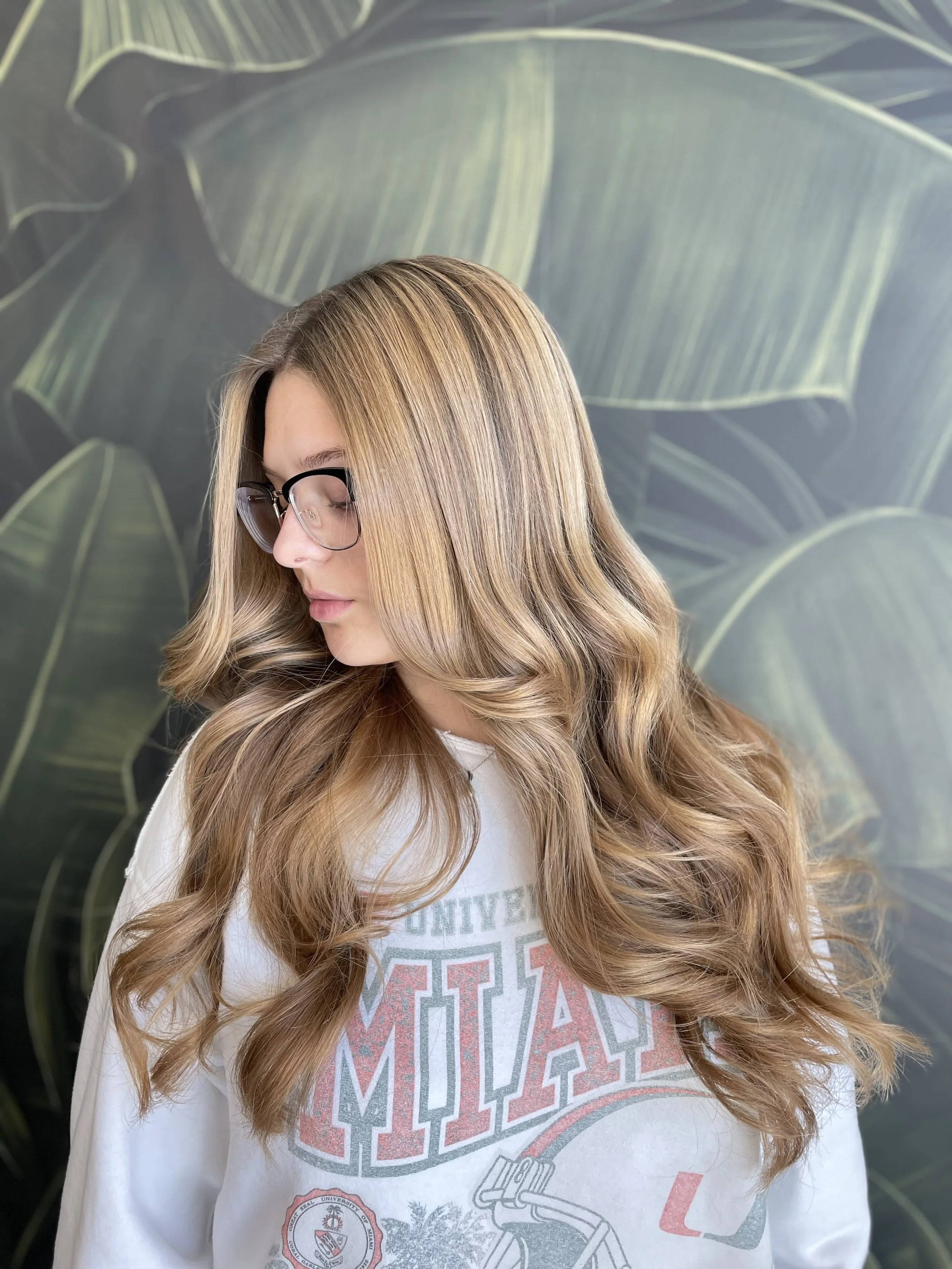 A woman with long, wavy, blonde hair and black eyeglasses looking down, standing in front of a backdrop with large green leaves.