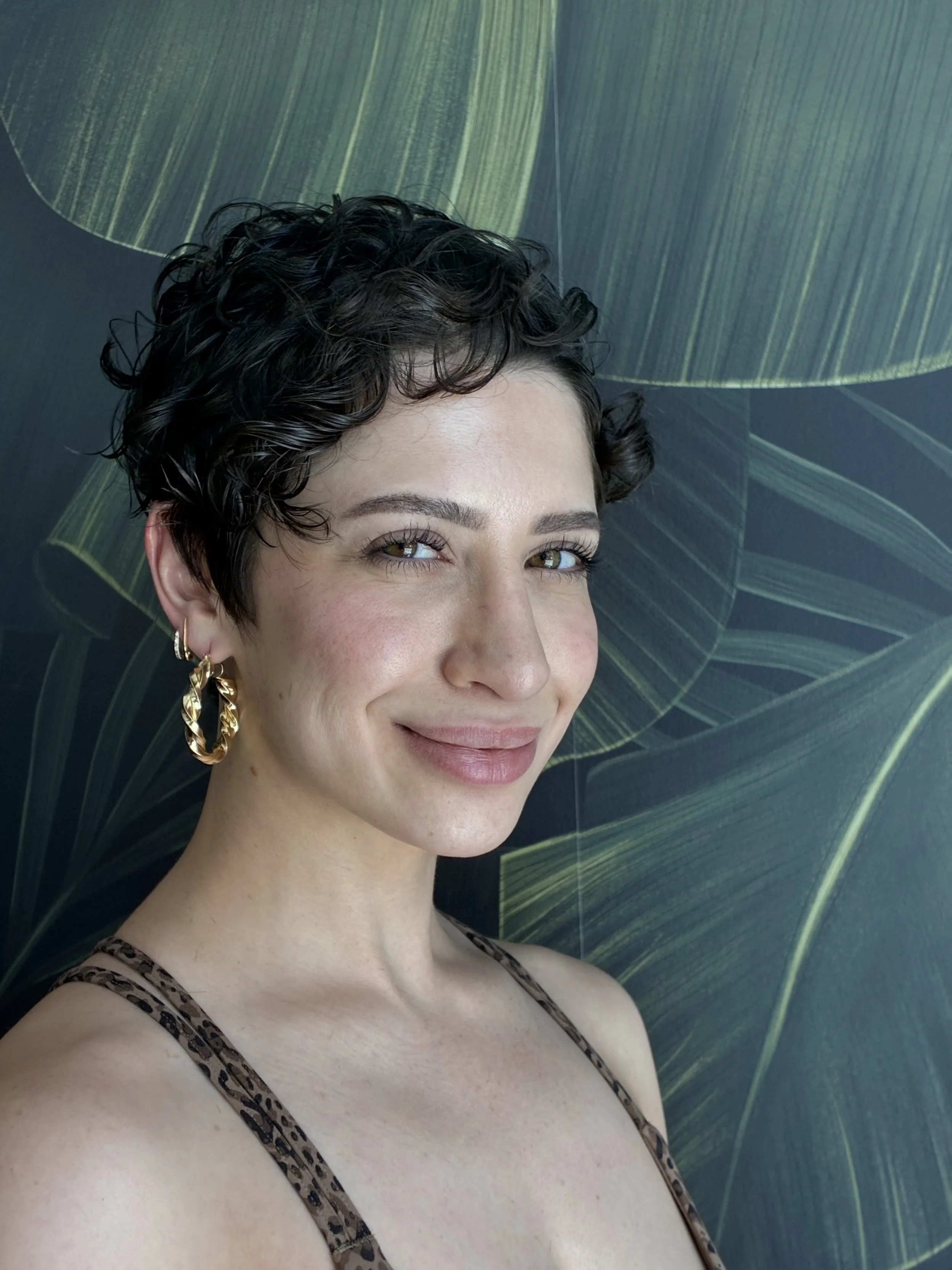 A woman with short, curly dark hair smiling in front of a green leaf-patterned background. She is wearing gold hoop earrings and a leopard print sleeveless top.