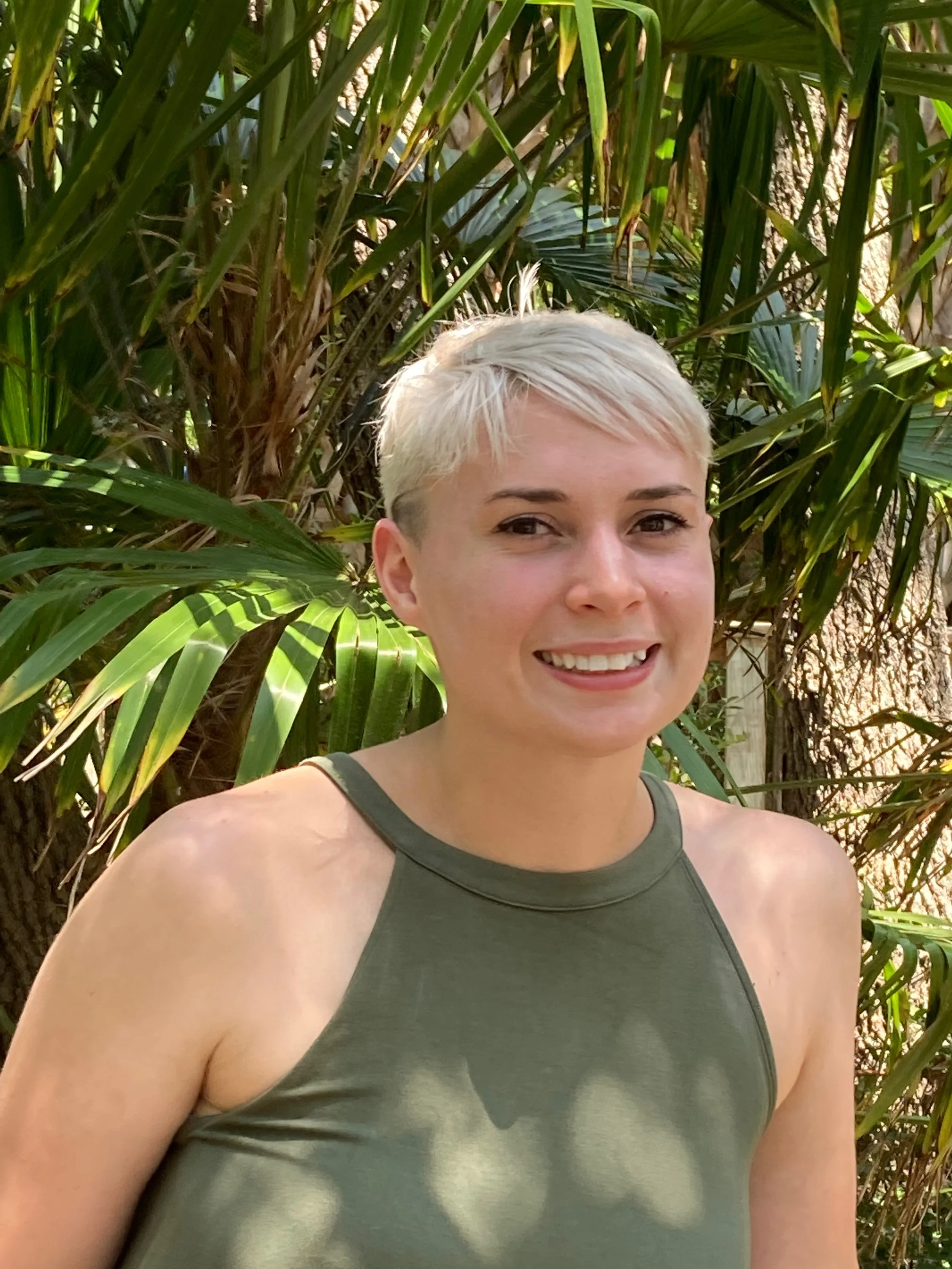 A woman with short blonde hair and a green sleeveless top smiling in front of tropical palm leaves.