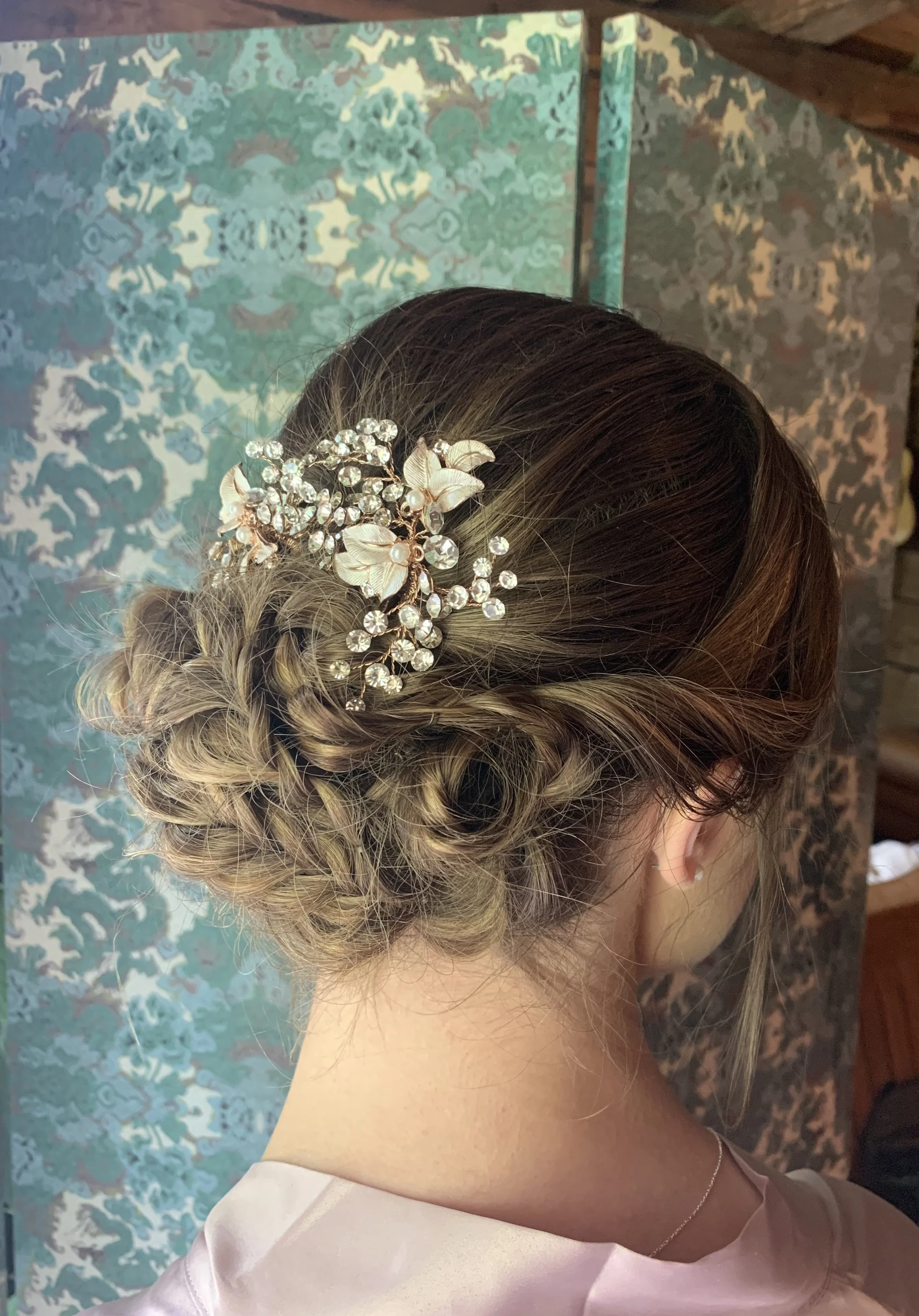 Side view of a woman's hairstyle with a decorative hairpiece of pearls, crystals, and floral accents, styled in an elegant updo with blonde and brown hair, set against a patterned background.