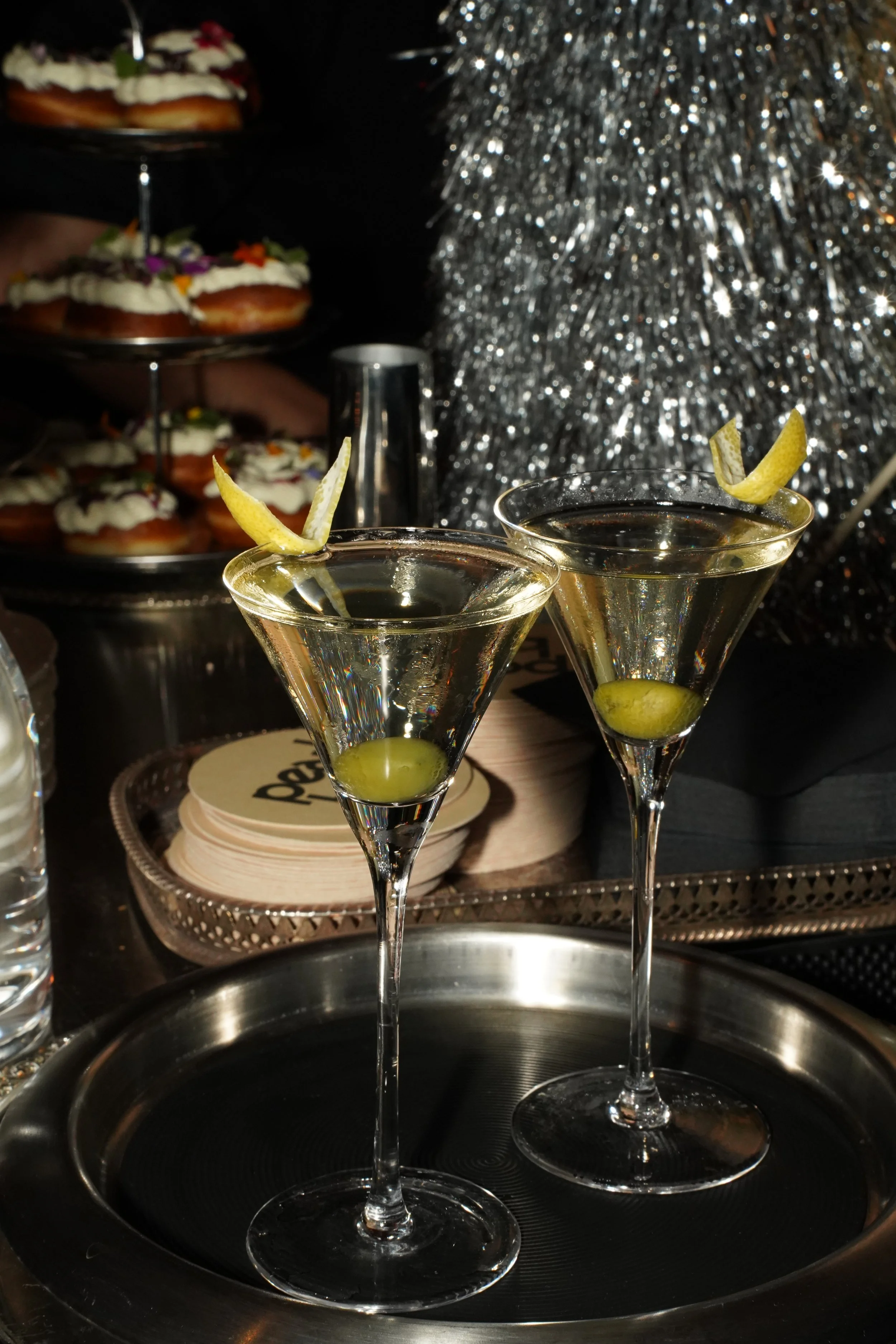 Two cocktails with lemon twists garnish in front of a display of donuts topped with cream and flowers, and a large silver tinsel decoration in the background.