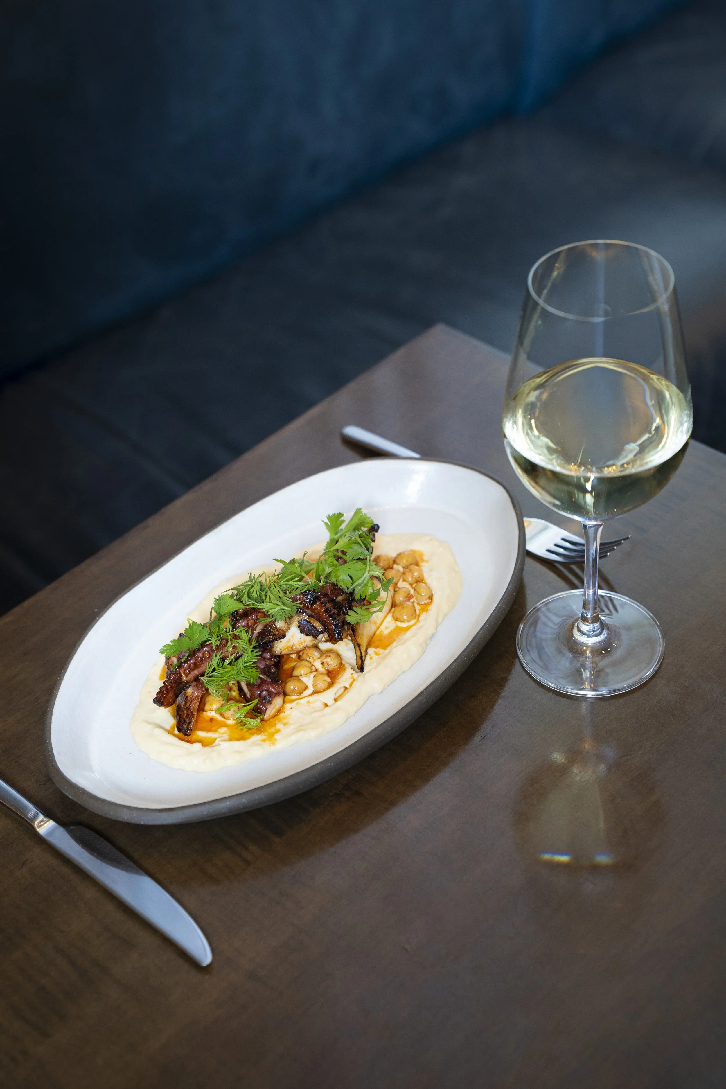 A plate of grilled octopus tentacles with herbs on a bed of creamy mashed potatoes accompanied by a glass of white wine on a wooden table.