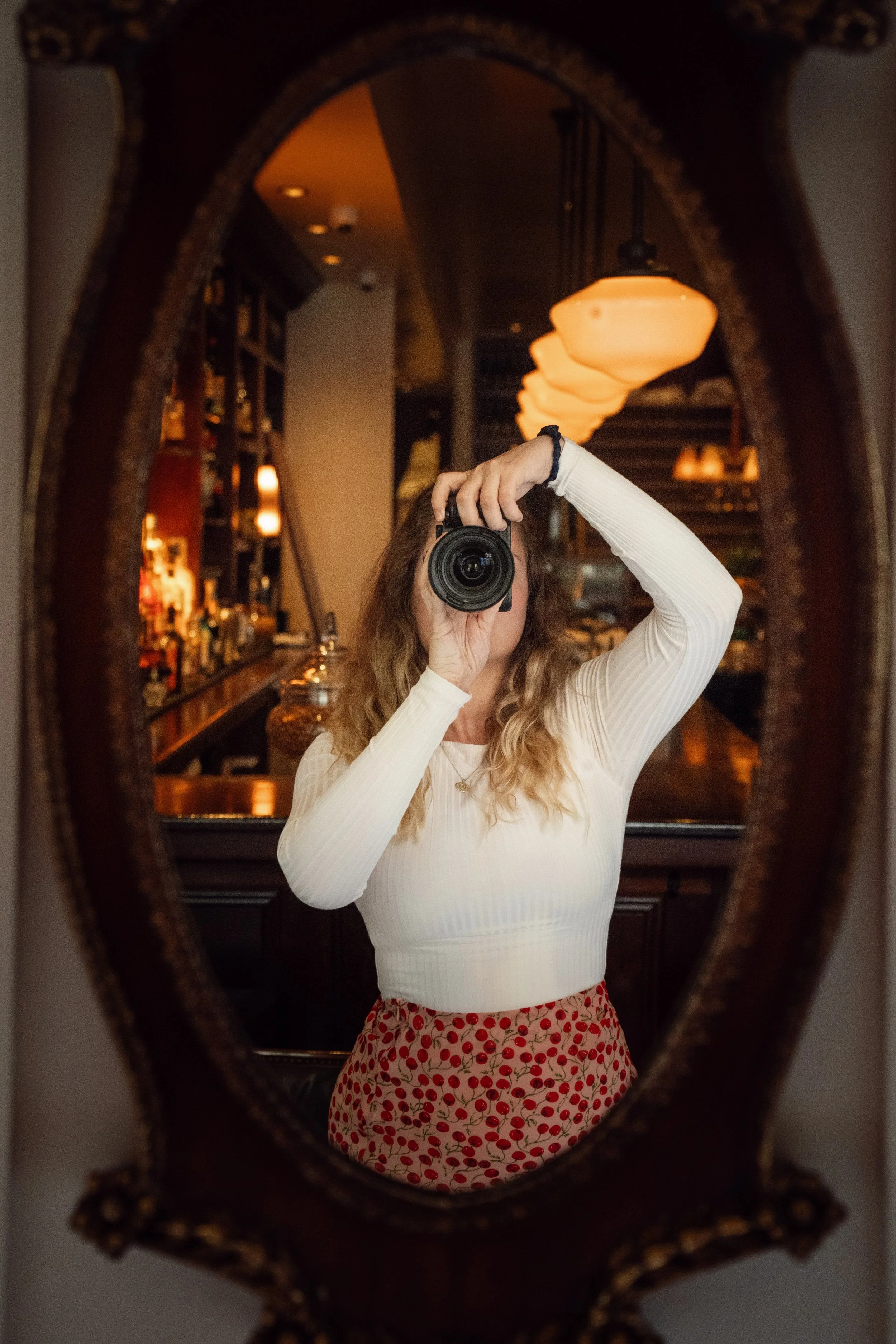 A woman taking a photo of herself in a mirror that is framed with an ornate, vintage-style border. Behind her, there is a bar with bottles and warm lighting fixtures hanging from the ceiling.