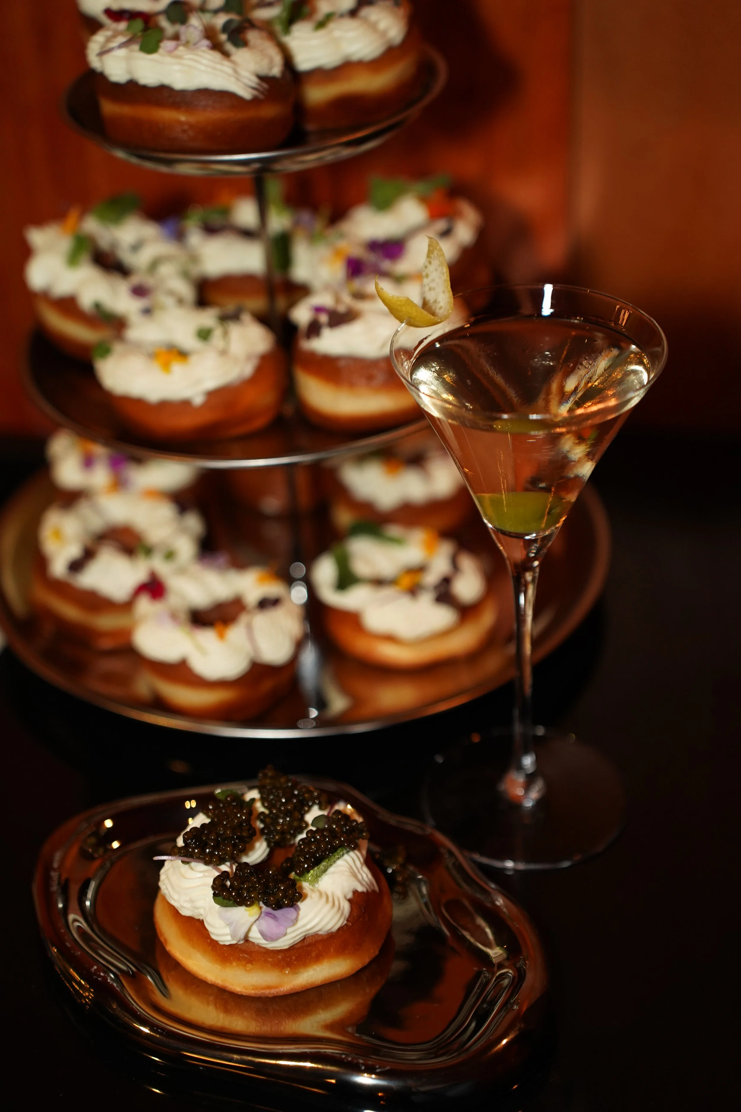 Assorted gourmet appetizers on glass plates, including a pastry topped with cream and caviar, a dessert tart with cream, chocolate, and edible flowers, and a cocktail with a lemon peel garnish, set against a dark background.