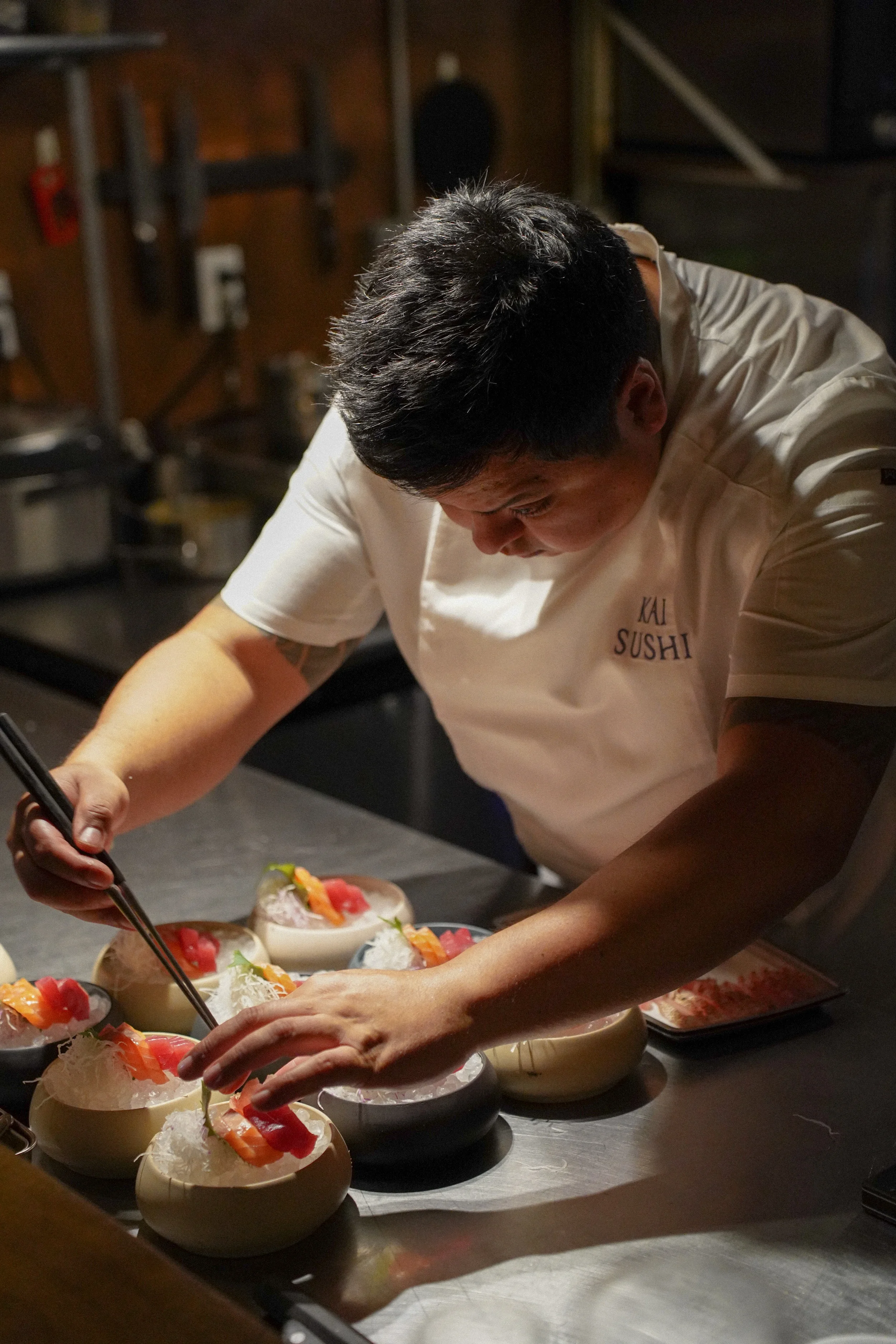 A chef is preparing colorful sushi bowls in a dimly lit kitchen, wearing a white shirt with the words 'KAI SUSHI' on it.