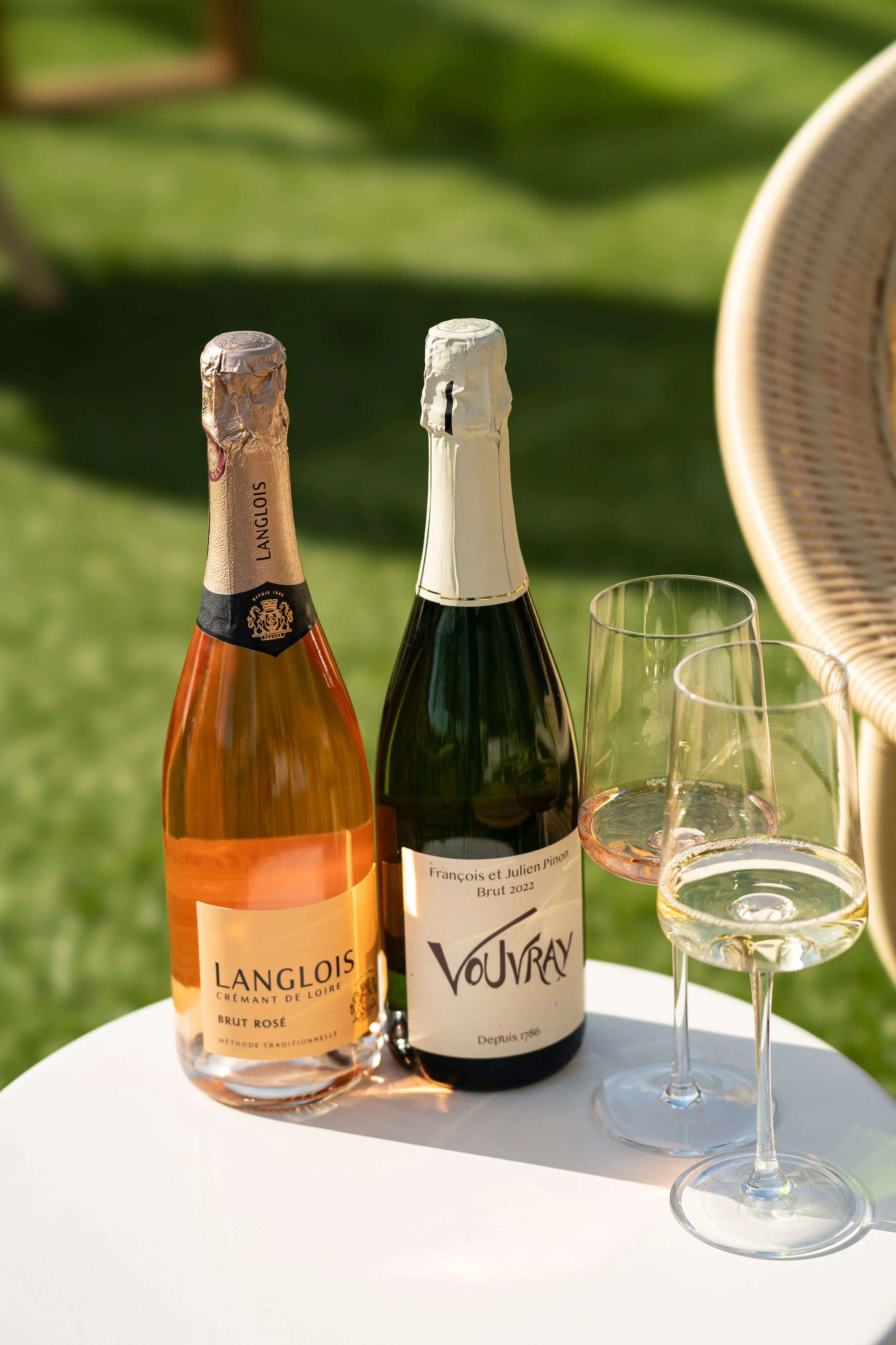 Two bottles of sparkling wine and a glass of white wine on a outdoor table with green foliage background.
