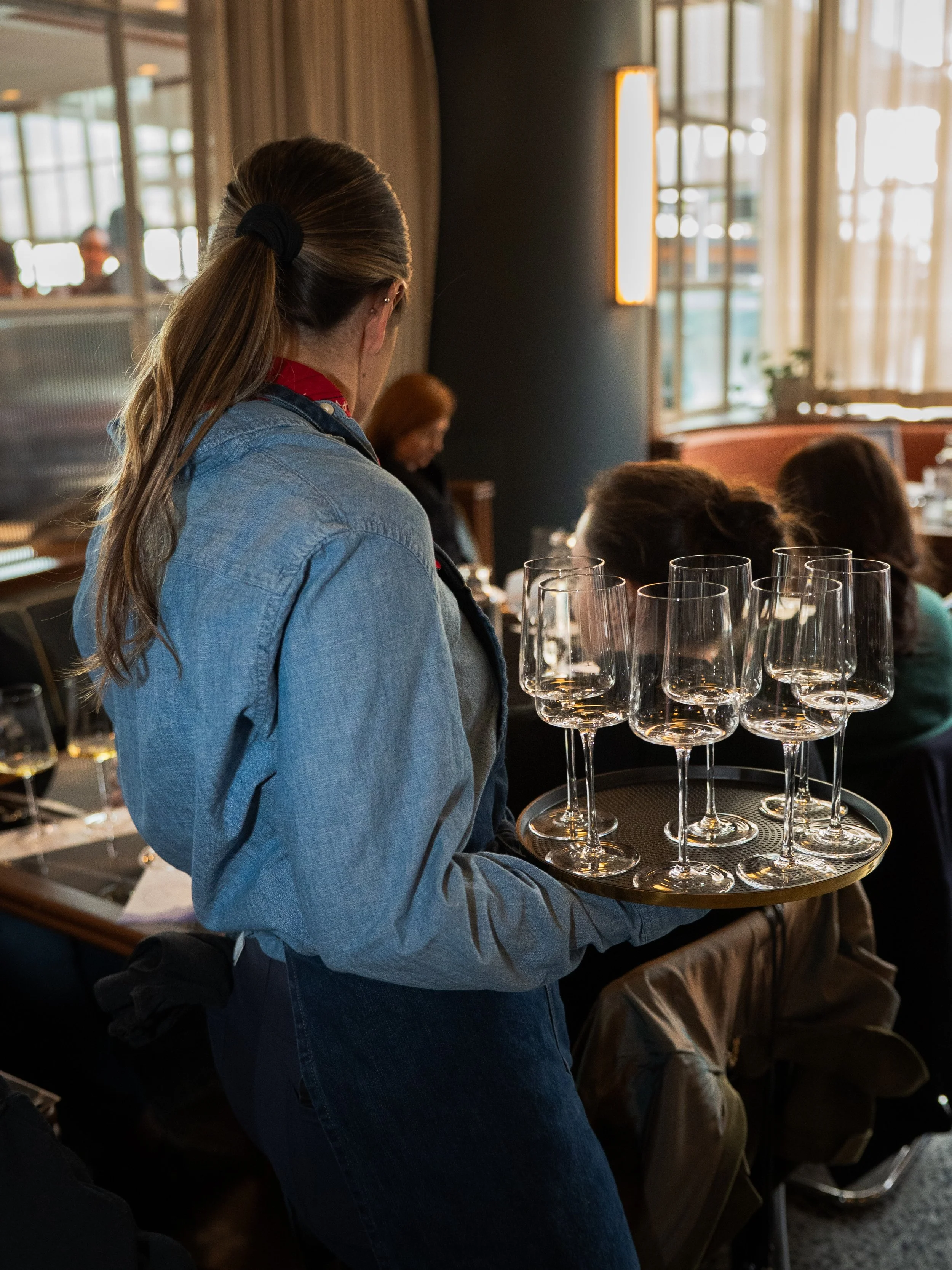 A woman in a denim jacket holds a tray with empty wine glasses in a restaurant or bar setting, with other patrons visible in the background.