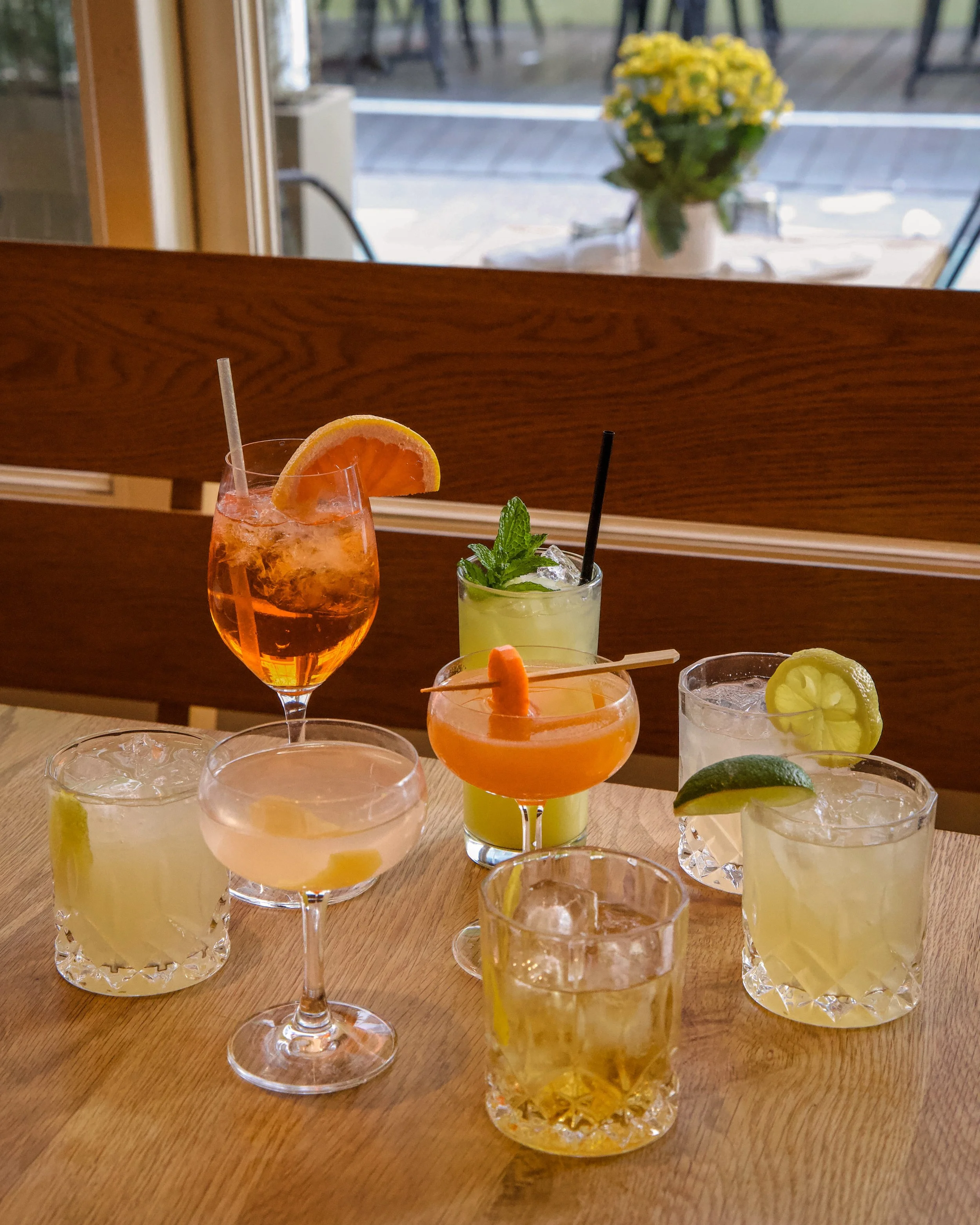 Various colorful cocktails with garnishes on a wooden table, with a background of a window and a flower vase outside.