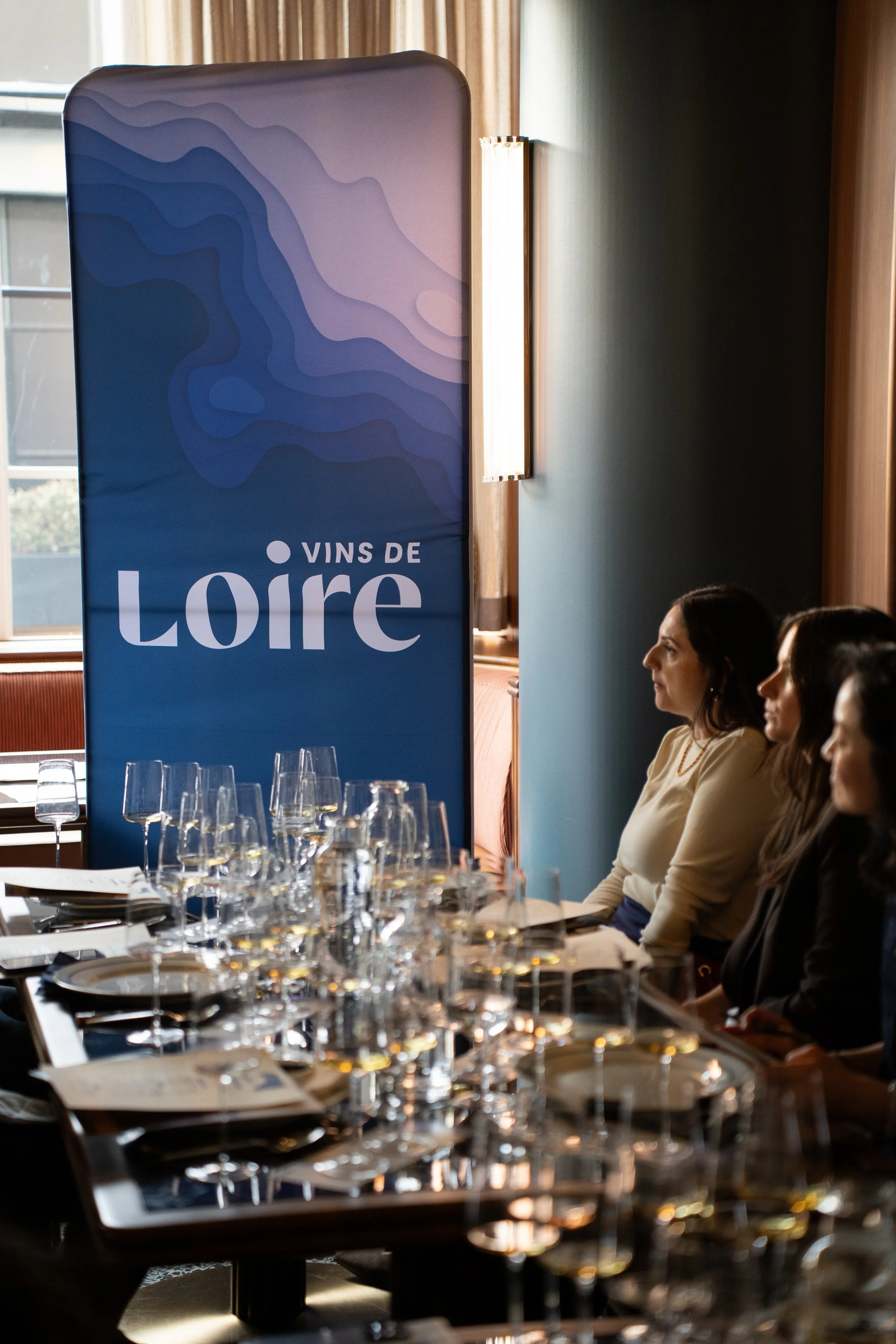 People sitting at a table set for a meal at an event called Vins de Loire, with wine glasses, plates, and utensils, in a room with natural light and wooden accents.
