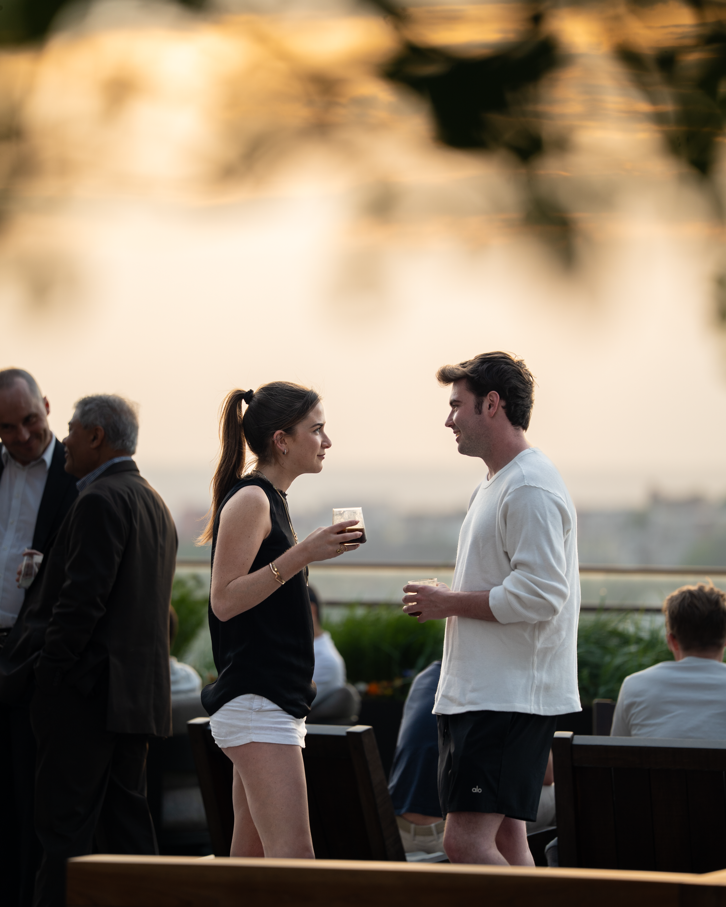 Two young adults, a woman with a ponytail holding a drink and a man in a white sweater, are facing each other and smiling outdoors during sunset. There are other people in the background, some in suits, and the setting appears to be a social gatherin