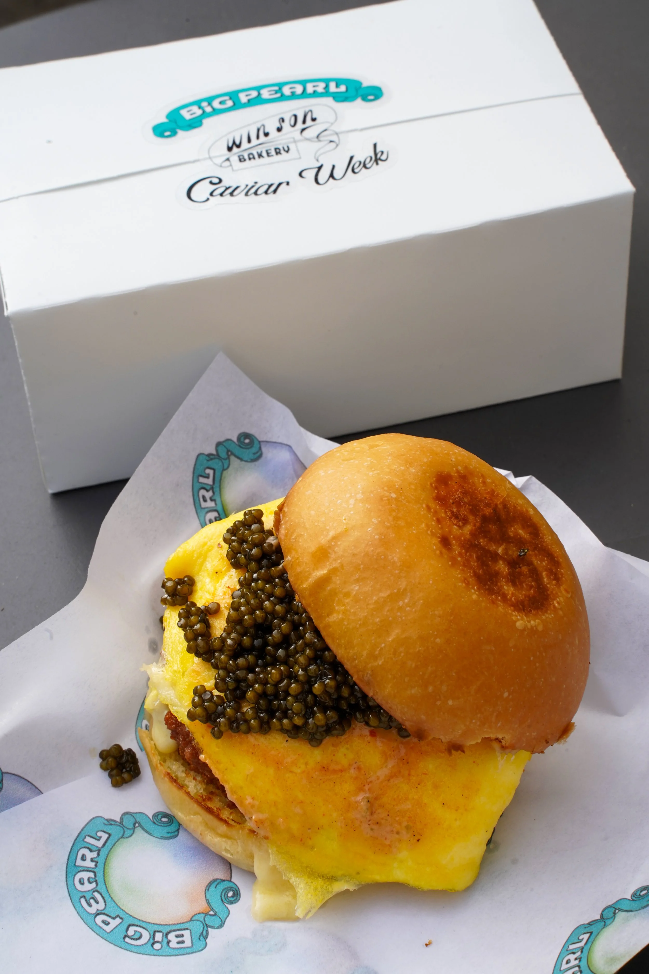 A sandwich with eggs, caviar, and a bun, placed on branded parchment paper, with a Big Pearl Bakery box in the background.
