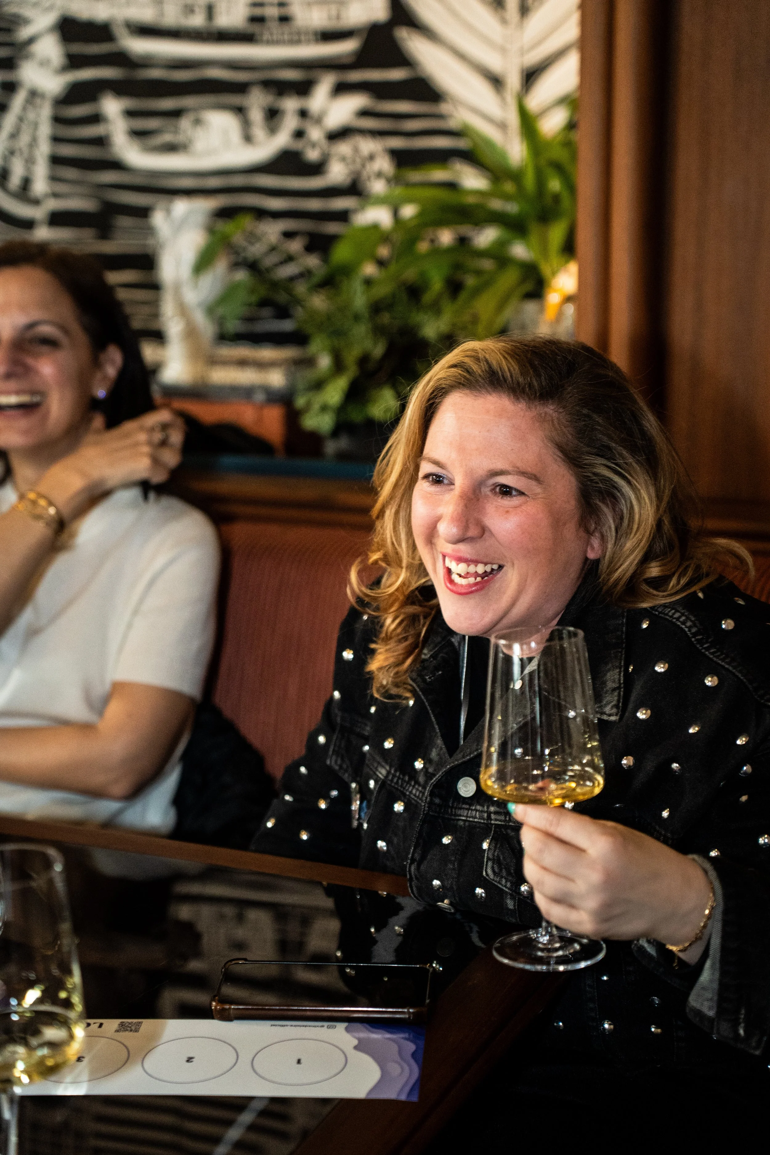 Two women enjoying a meal in a restaurant; one woman with curly hair holding a glass of white wine, smiling; the other woman with dark hair, also smiling; background features a black and white abstract wall art and a green plant.