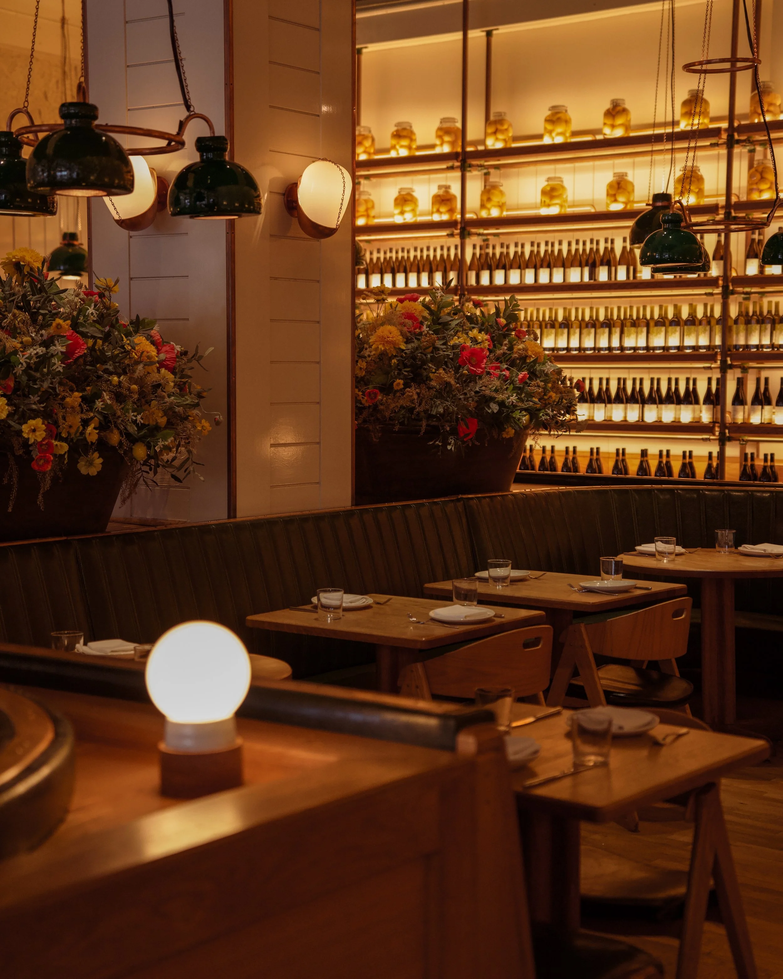 Interior of a cozy restaurant with wooden tables set with glasses and plates, soft warm lighting, plants with flowers on a shelf, and shelves filled with bottles and jars in the background.