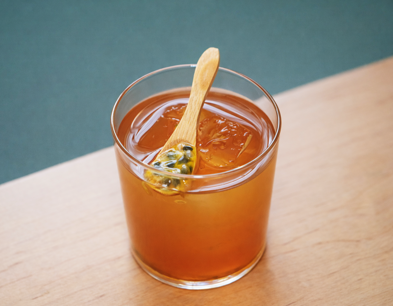A clear glass filled with amber-colored passion fruit iced tea with ice cubes and a wooden stir stick with passion fruit seeds inside.