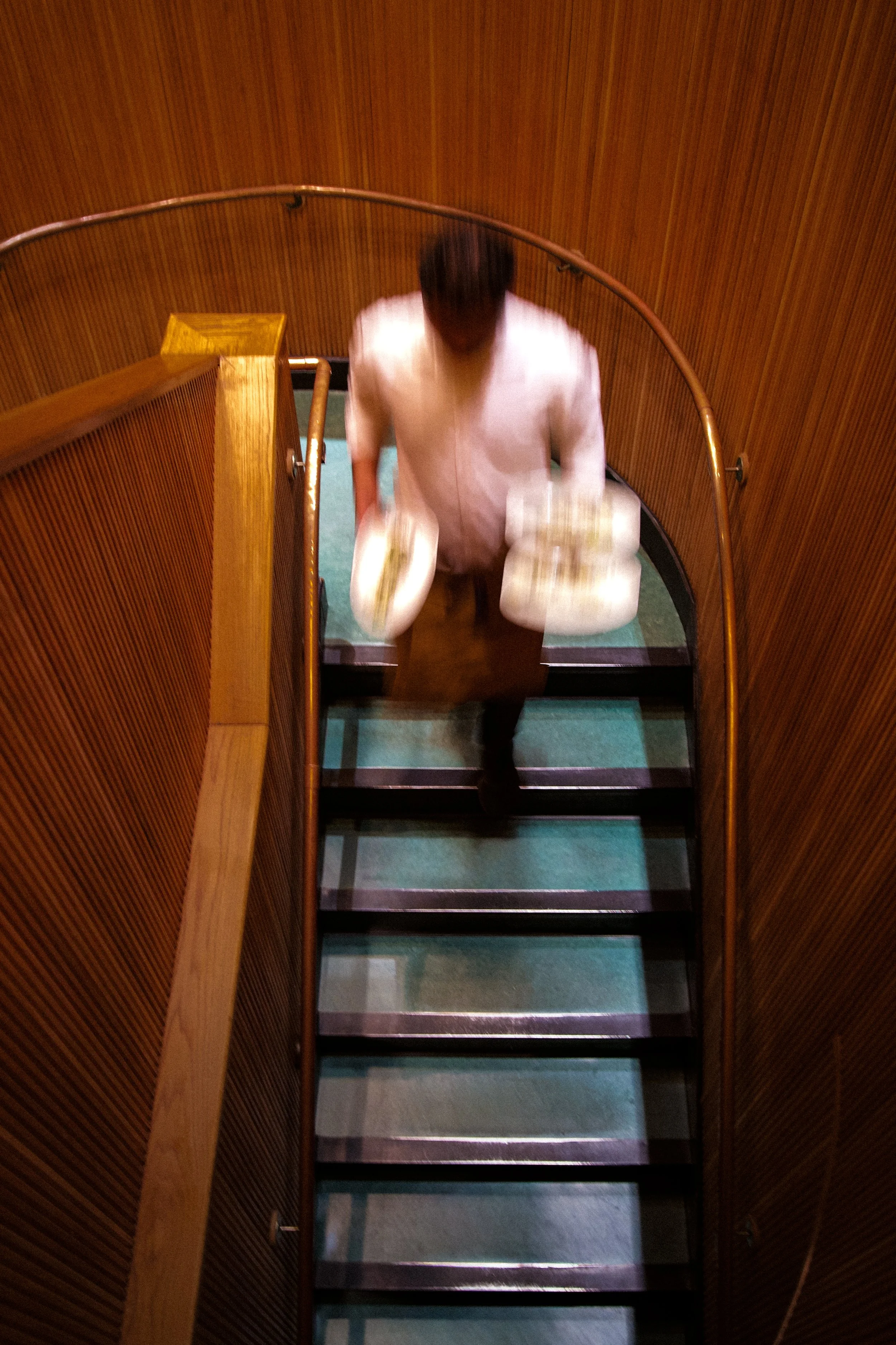 A person with dark hair wearing a white shirt and brown pants, carrying two trays of food while descending a narrow staircase with wooden walls and green carpeted steps.