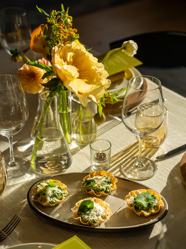 A dining table with a floral centerpiece, glassware, a small candle, and a plate of four savory tartlets garnished with green leaves and cheese.