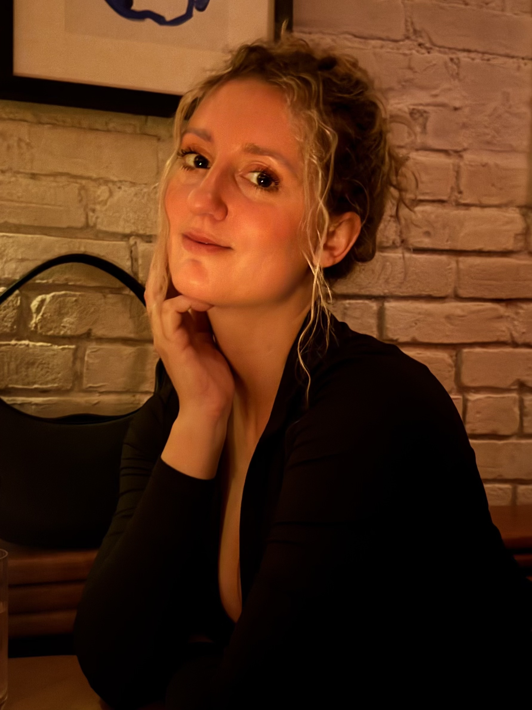 Young woman with curly blond hair sitting in a cozy indoor setting with a brick wall behind her, wearing a black top, resting her chin on her hand and looking into the camera with a gentle smile.