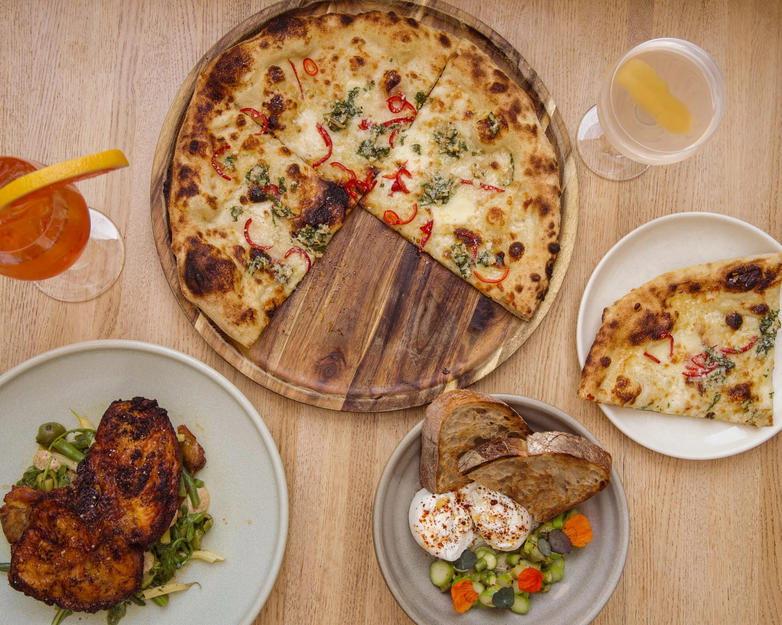 Various dishes including pizza with red peppers and greens, grilled chicken with greens, bread, and Mediterranean salads on plates and a wooden table.