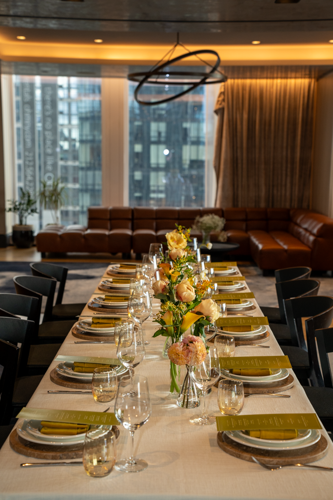 Elegant dining table set for a formal event with floral centerpieces, glassware, and yellow napkins, in a modern upscale restaurant or hotel banquet room.