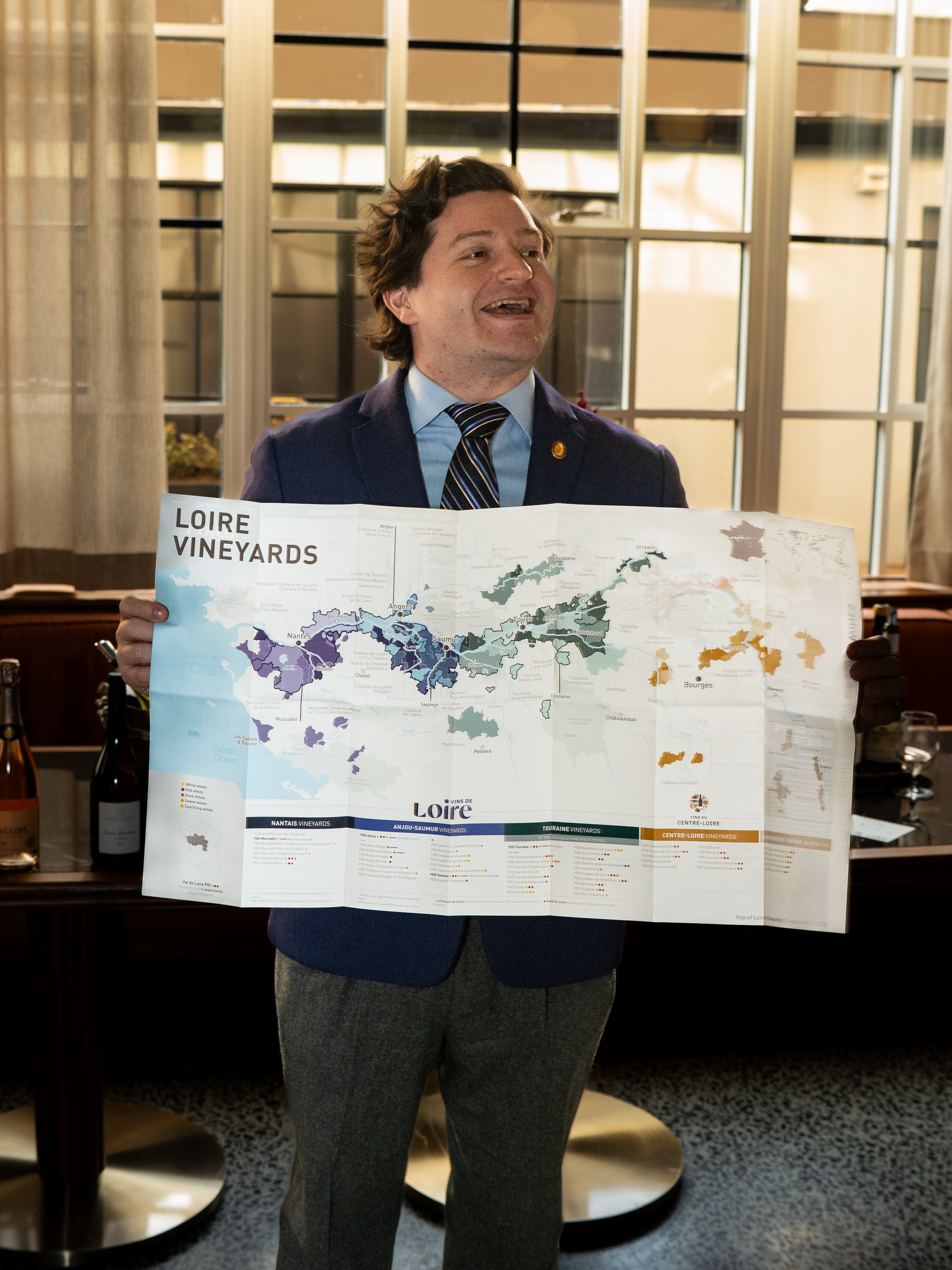 A man in a blue suit jacket and a striped tie holding a map of Loire vineyards, with a wooden furniture piece behind him and a window with beige curtains in the background.