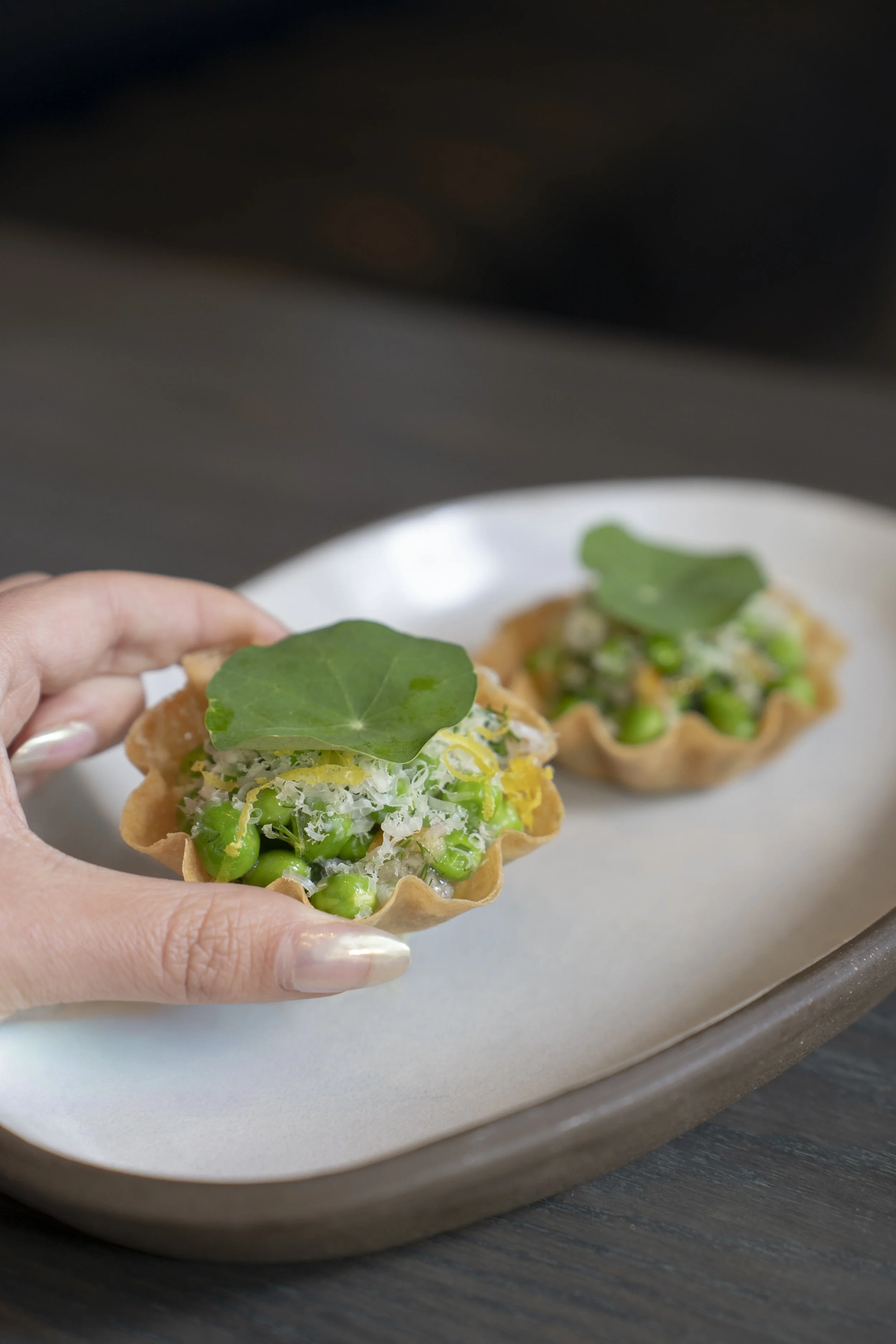 Close-up of a hand holding a small tart filled with green peas, grated cheese, and topped with a green leaf, with another similar tart on a white plate in the background.