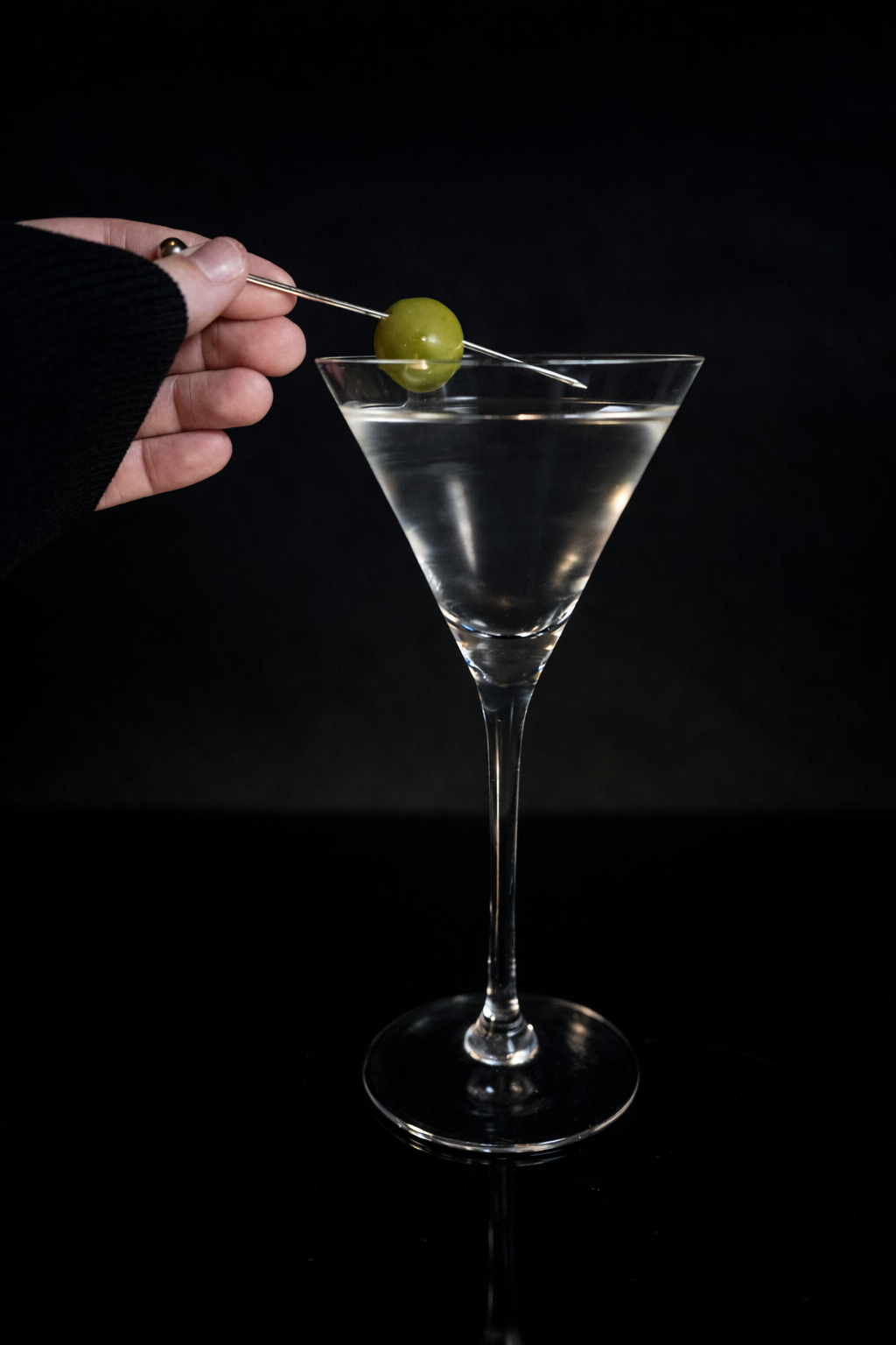 Person holding green olive with a cocktail pick over a martini glass filled with clear liquid against a black background.