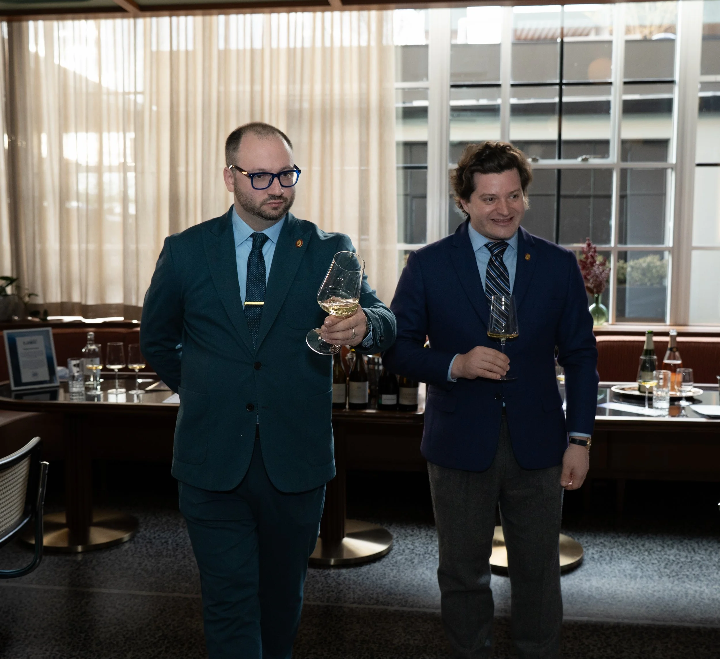 Two men dressed in suits standing in a room with large windows and curtains, holding wine glasses, engaged in conversation.