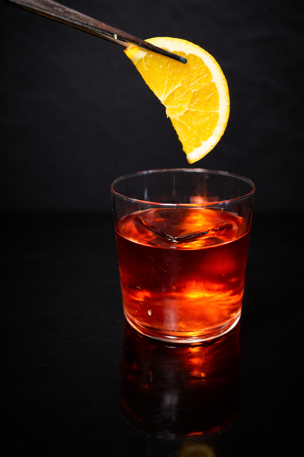 A close-up of a glass of iced red beverage with a lemon wedge held above it by a pair of dark chopsticks against a black background.