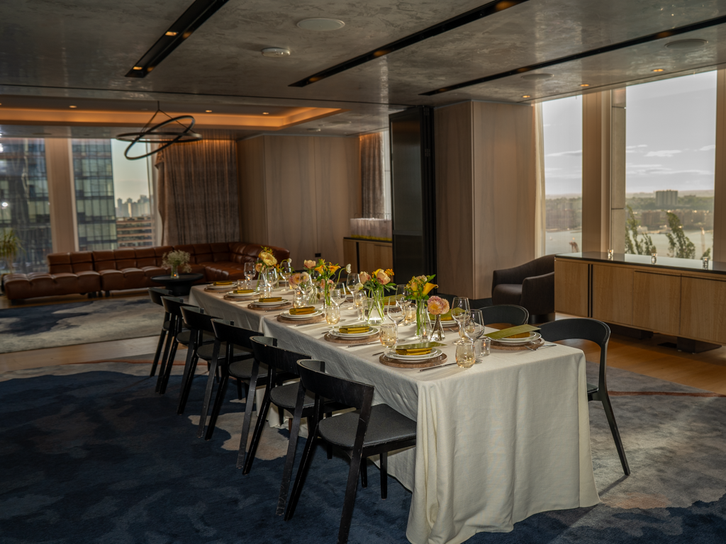 Elegant dining room with a long table set for a meal, featuring flowers in vases, wine glasses, and plates, overlooking cityscape through large windows.