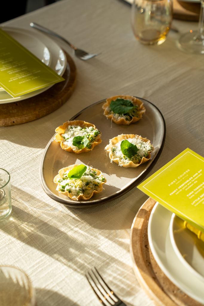 Four small tartlets with cheese and green garnish on a gray oval plate, on a table with plates, utensils, and drinks.