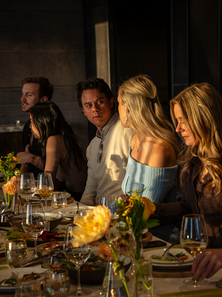 People sitting at a dinner table with wine glasses, flowers, and dishes, engaged in conversation.