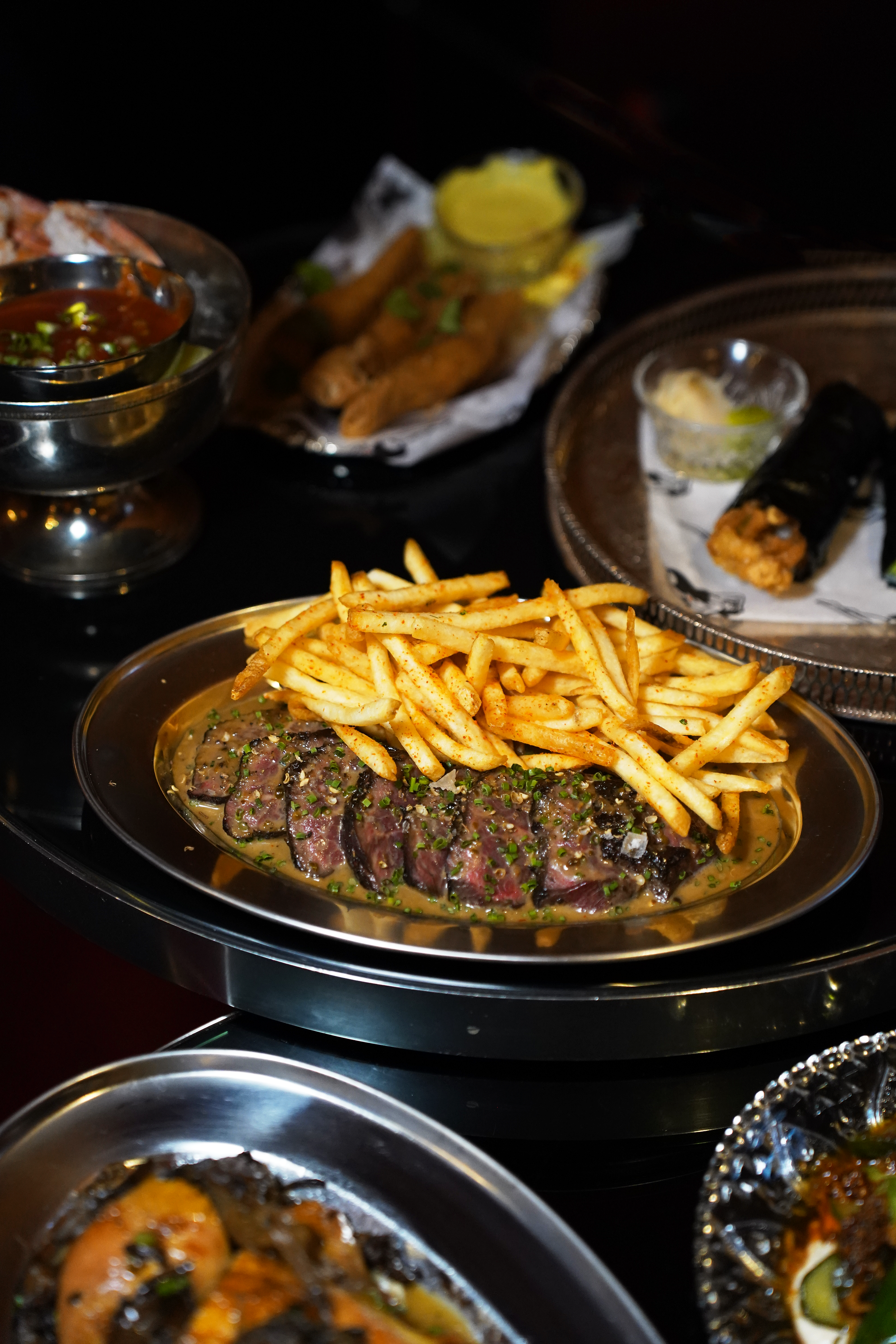 A variety of dishes including steak with fries, spring rolls with a lemon wedge, and sushi rolls, served on a dark table.