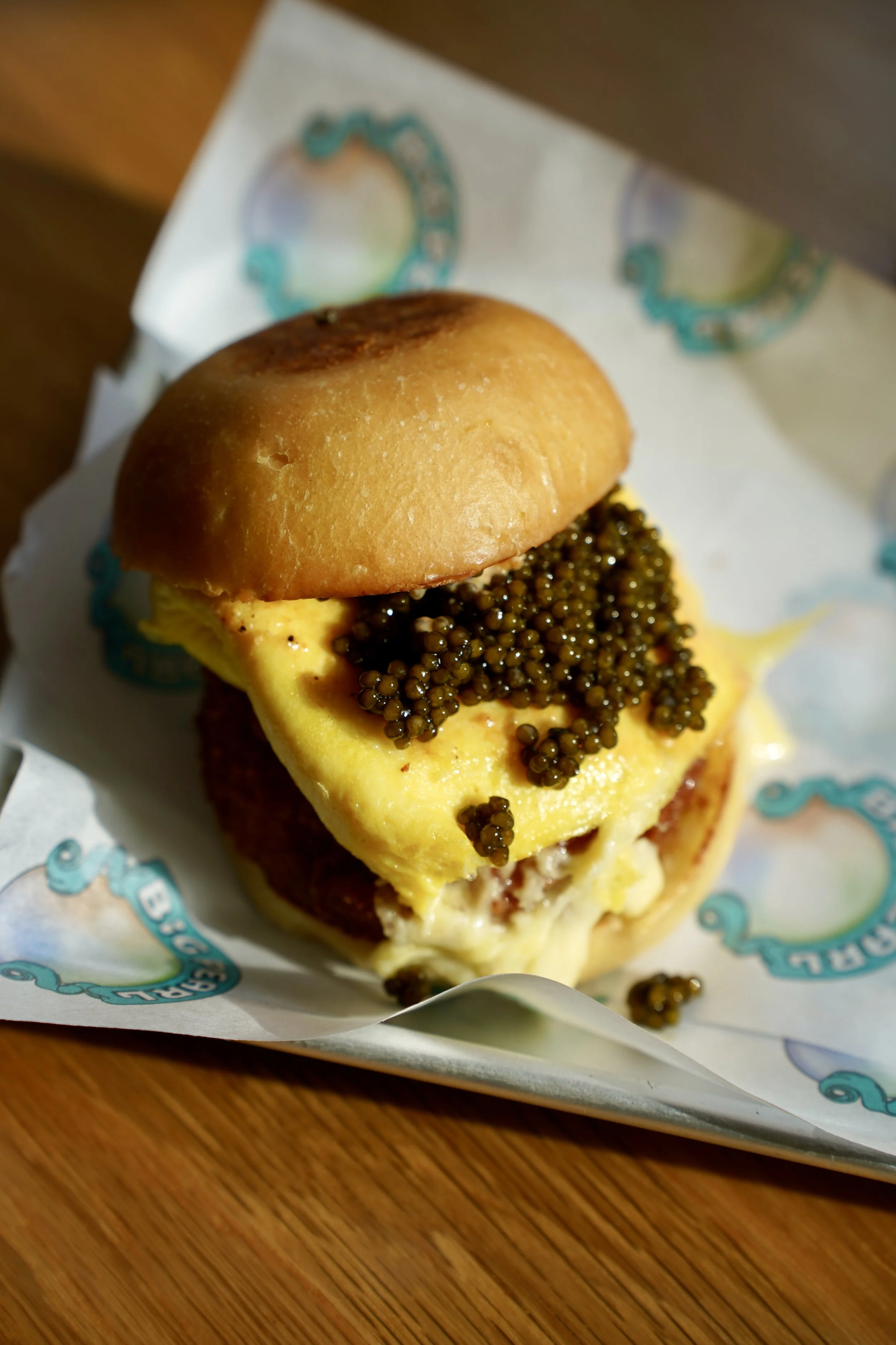 Close-up of a breakfast sandwich with scrambled eggs, black caviar, and a toasted bun, placed on branded parchment paper on a wooden surface.