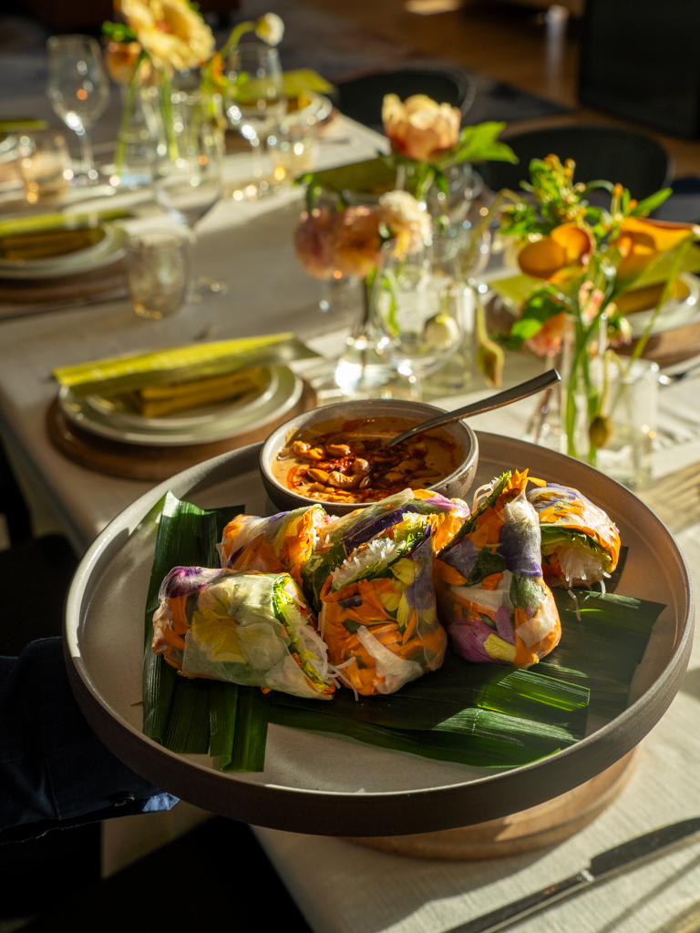 Spring roll appetizers with dipping sauce on a round platter, set on a decorated dining table with floral arrangements and dinnerware.