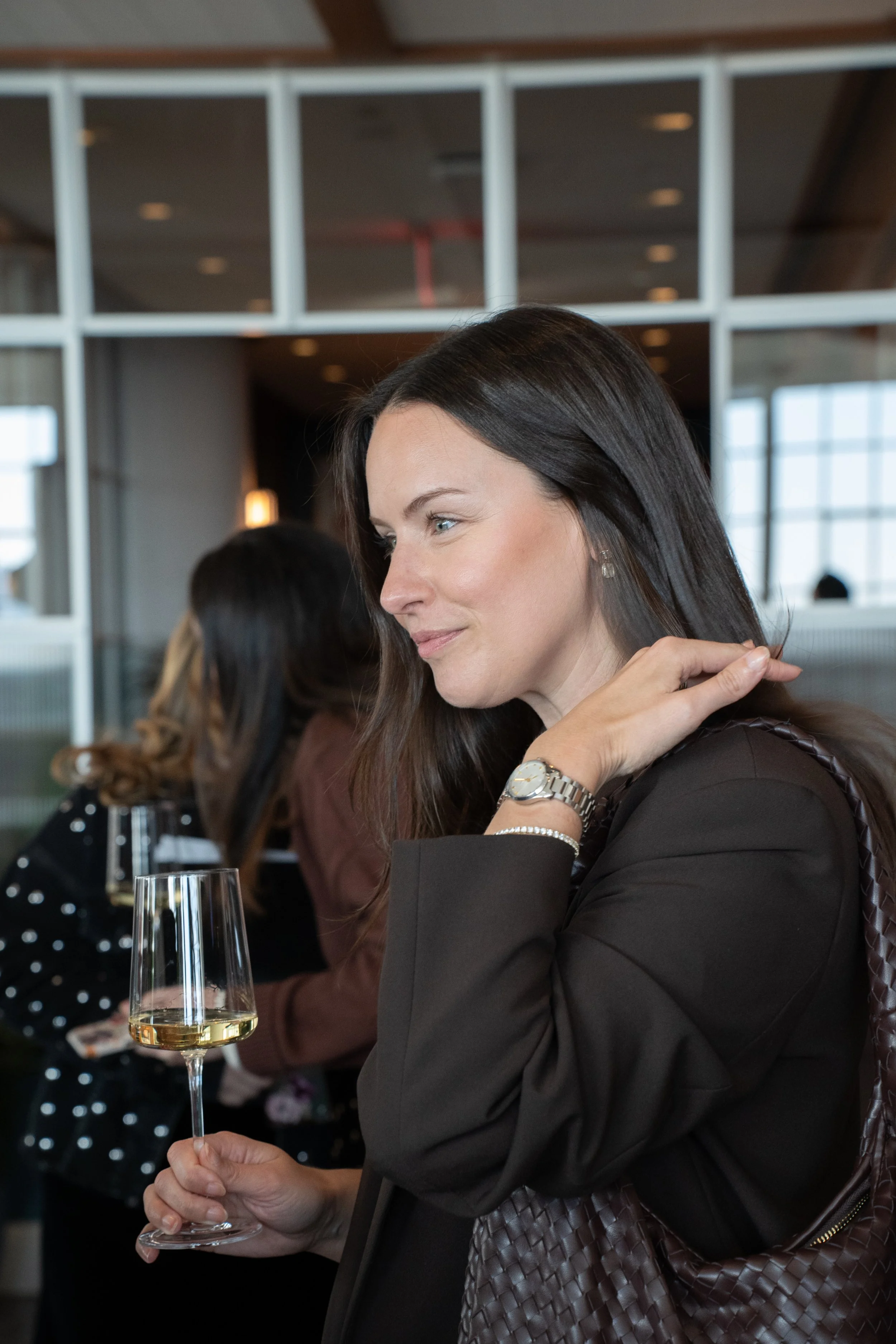 A woman with dark hair wearing a black blazer, silver watch, and earrings, holding a glass of white wine at a social gathering in a modern indoor space.