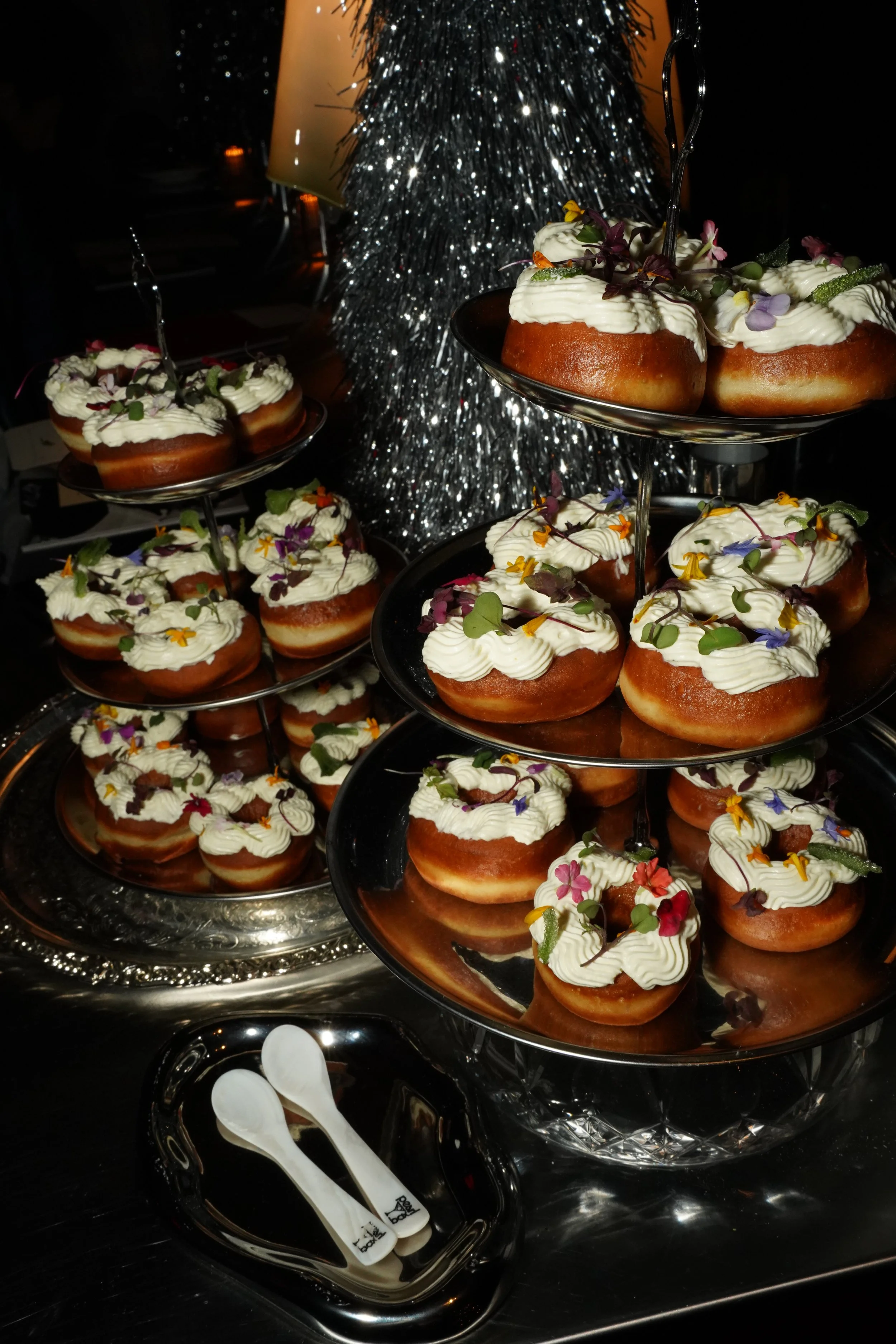 A multi-tiered display of decorated doughnuts with white icing and colorful edible flowers, set on silver trays with white plastic utensils nearby, in front of a shiny silver tinsel Christmas tree