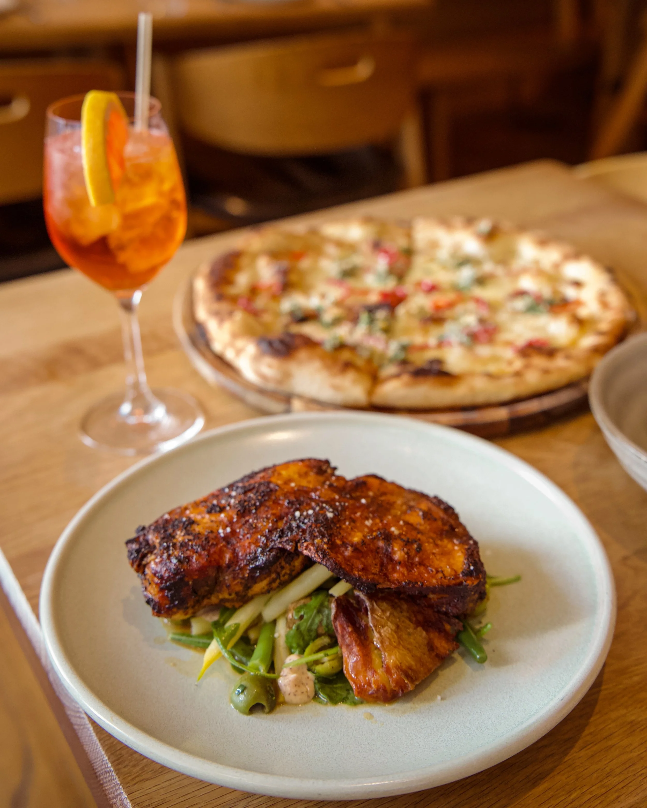A plate with a piece of grilled chicken on a bed of greens and roasted vegetables, with a glass of iced tea garnished with a lemon wedge and a straw in the background, along with a pizza on another dish.