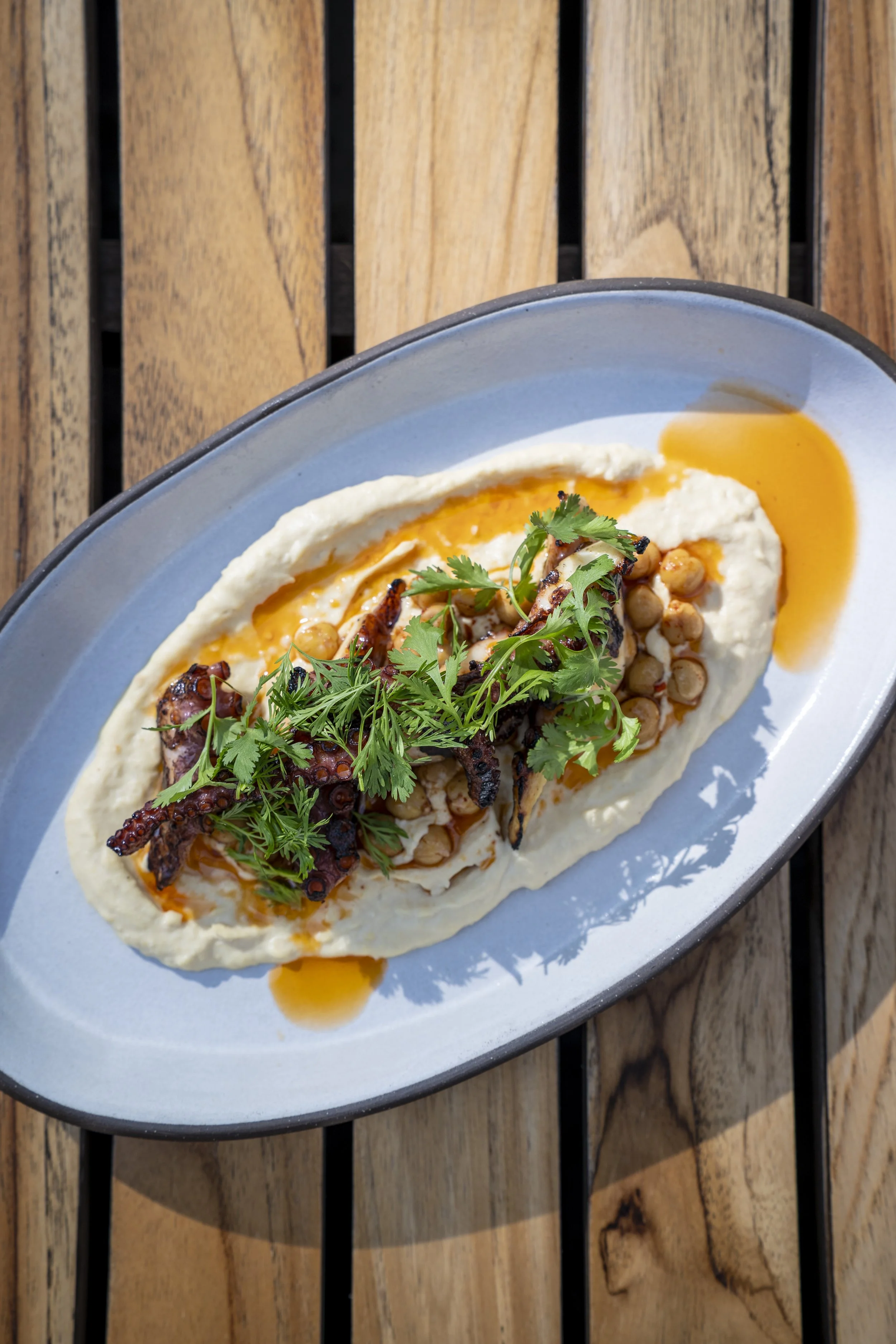 A plate of hummus topped with grilled octopus, chickpeas, herbs, and drizzled with oil, served on a ceramic plate placed on a wooden table.
